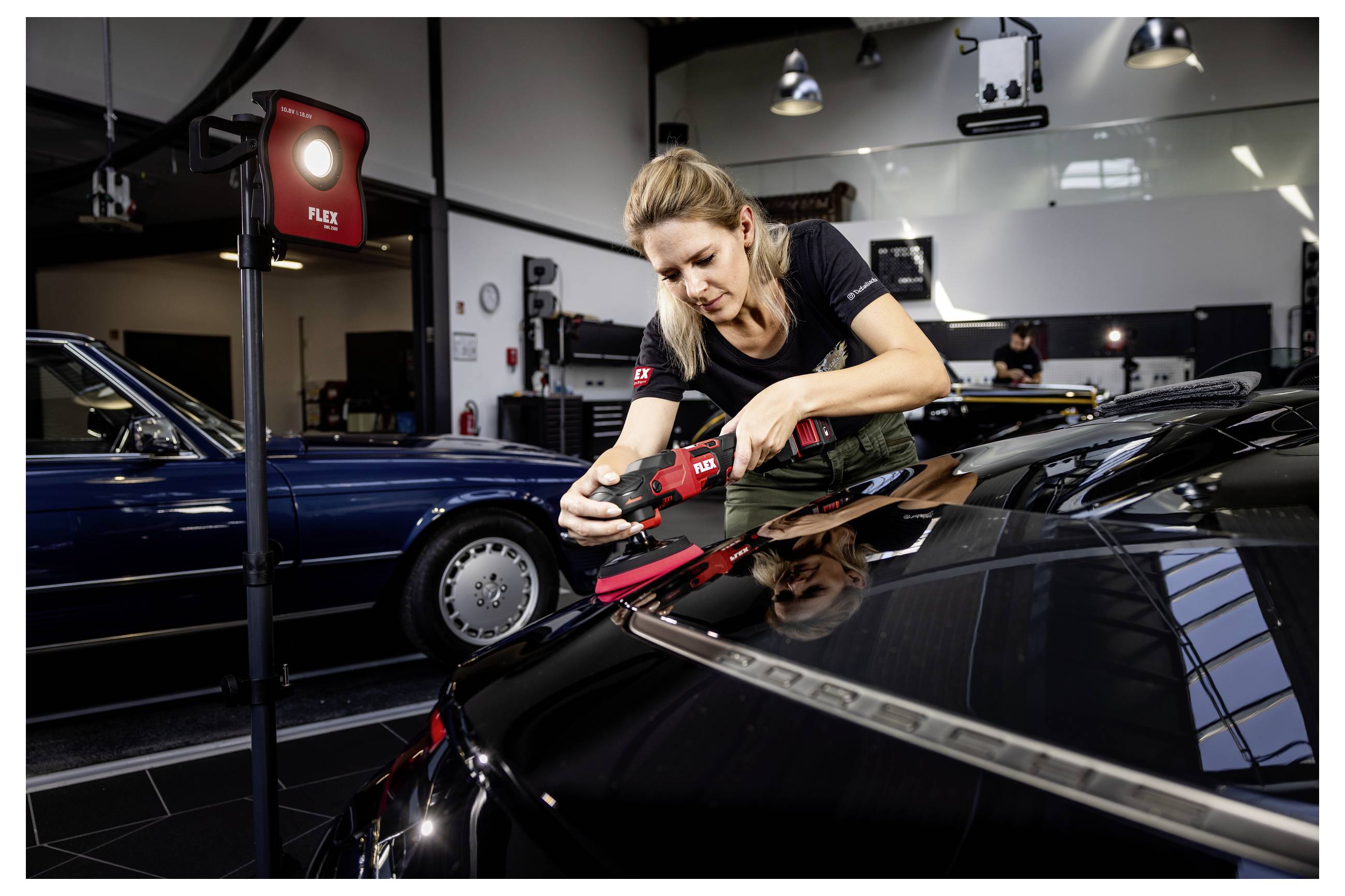 Žena používající elektrickoslouč na kapotě černého auta v dílně pro údržbu automobilů, s dalším klasickým autem v pozadí.