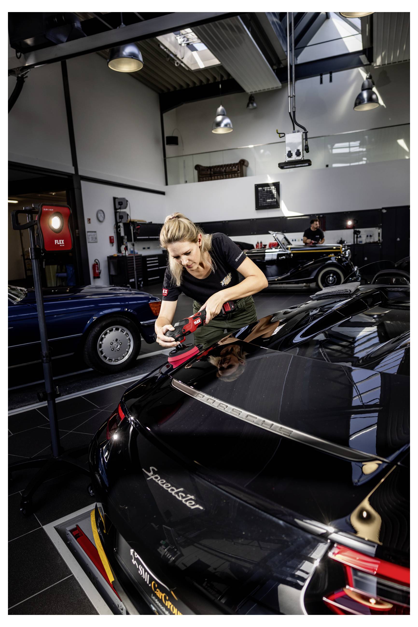 Žena leštící černý Porsche Speedster v autosalónu, v pozadí další klasické automobily.