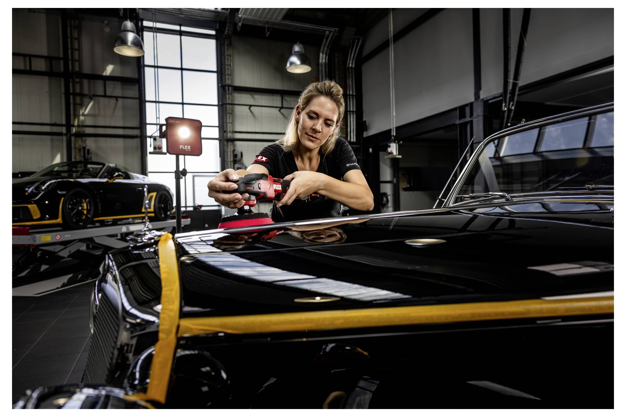 Žena leštící kapotu černého sportovního vozu v garáži, s dalším luxusním automobilem v pozadí.