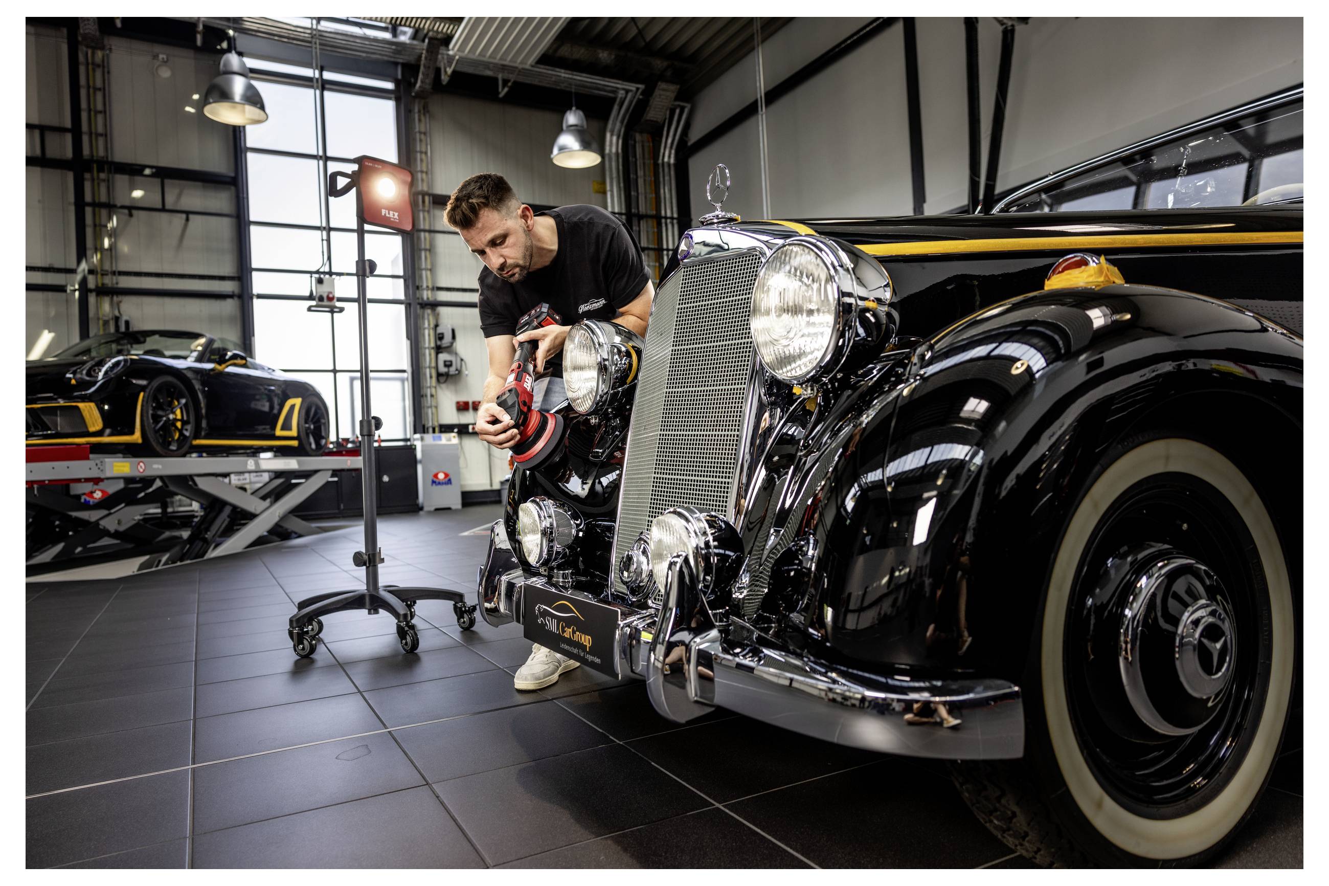 Mechanik leští reflektor černého veteránského automobilu v garáži, v pozadí je další klasický vůz na zvedáku.