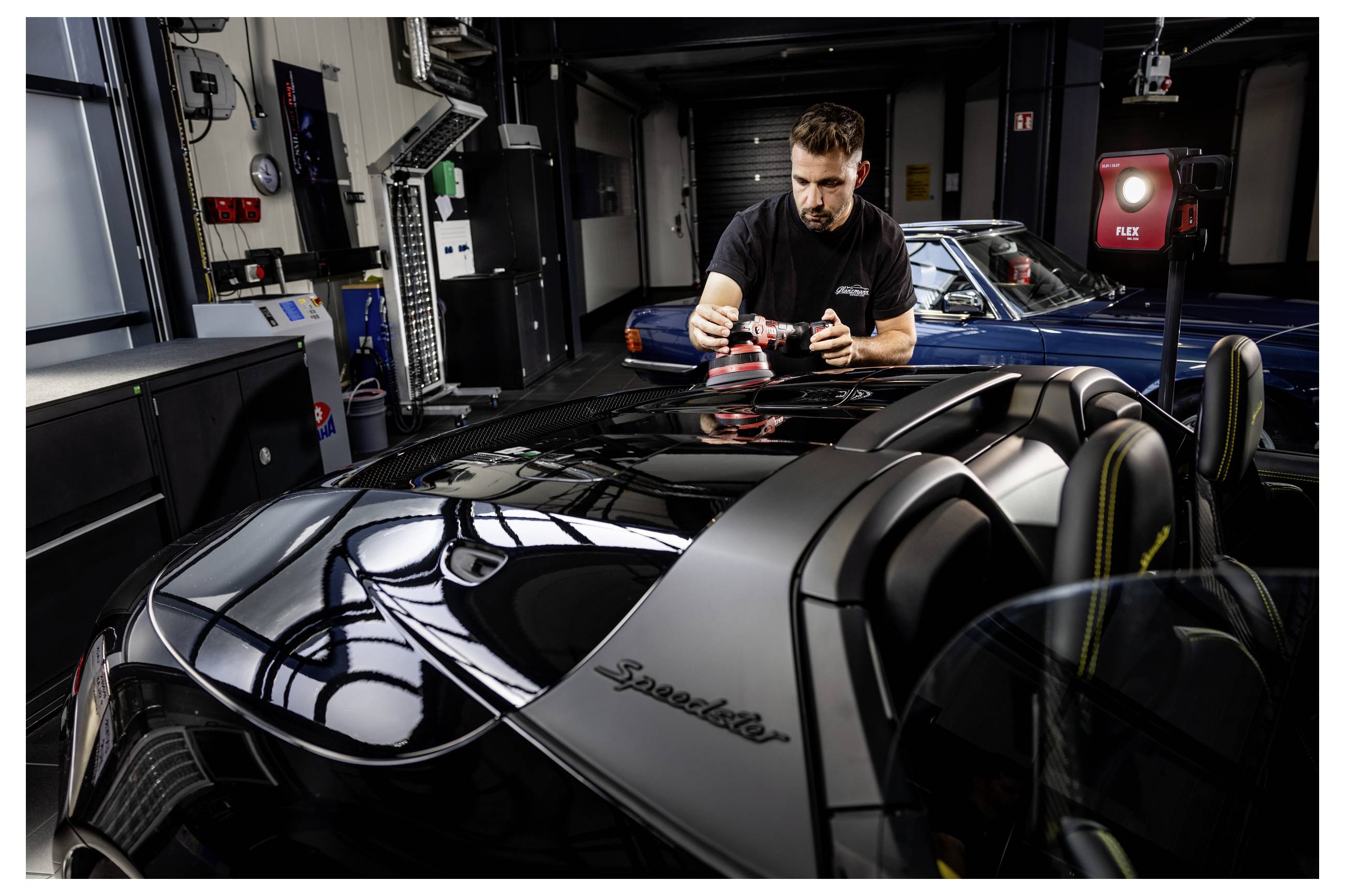 Muž kontrolující lesklou černou střechu Lamborghini Speedster v garáži s modrým klasickým autem v pozadí.