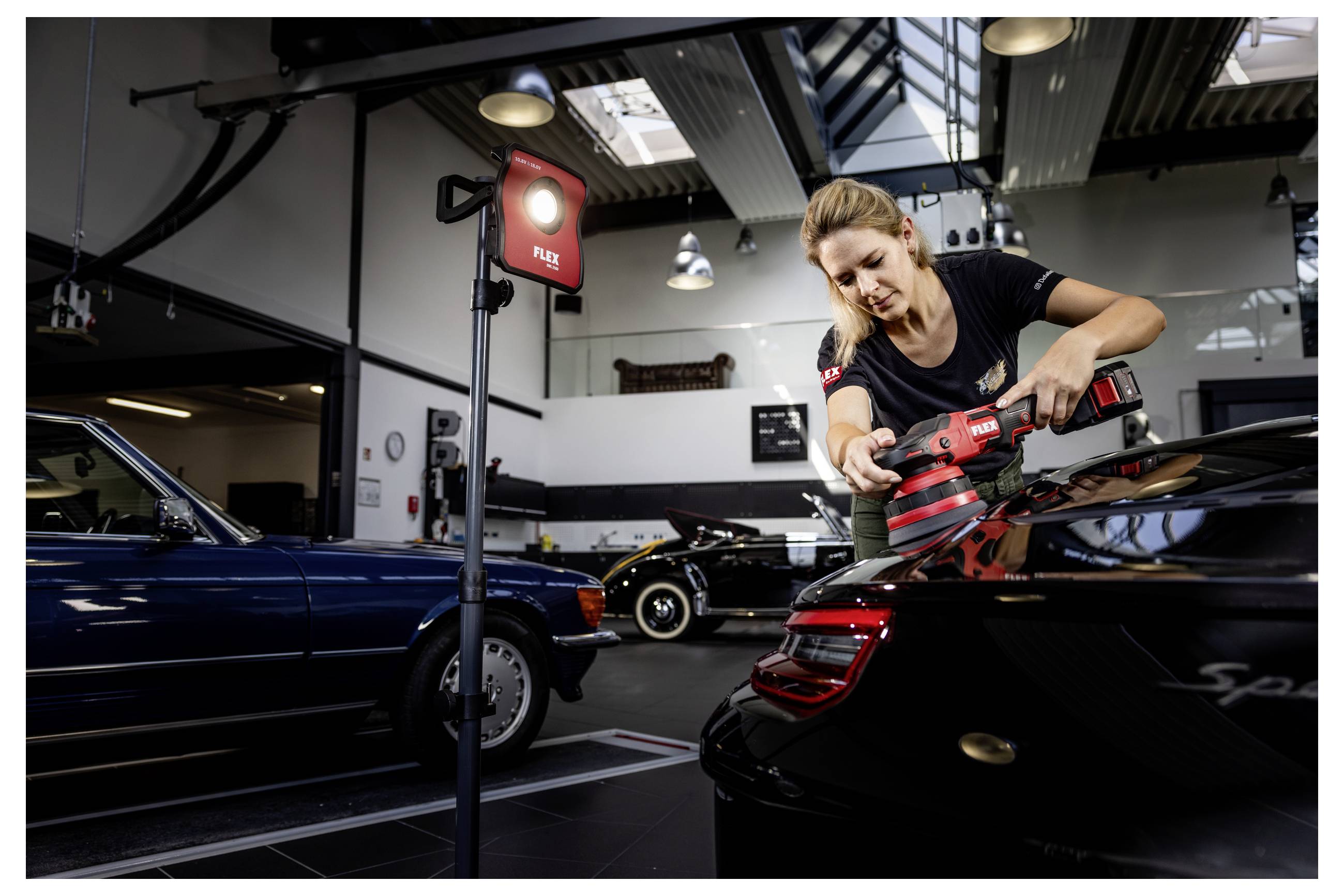 Žena leštící zadní část černého sportovního vozu Porsche v autodílně, s dalšími klasickými auty v pozadí.