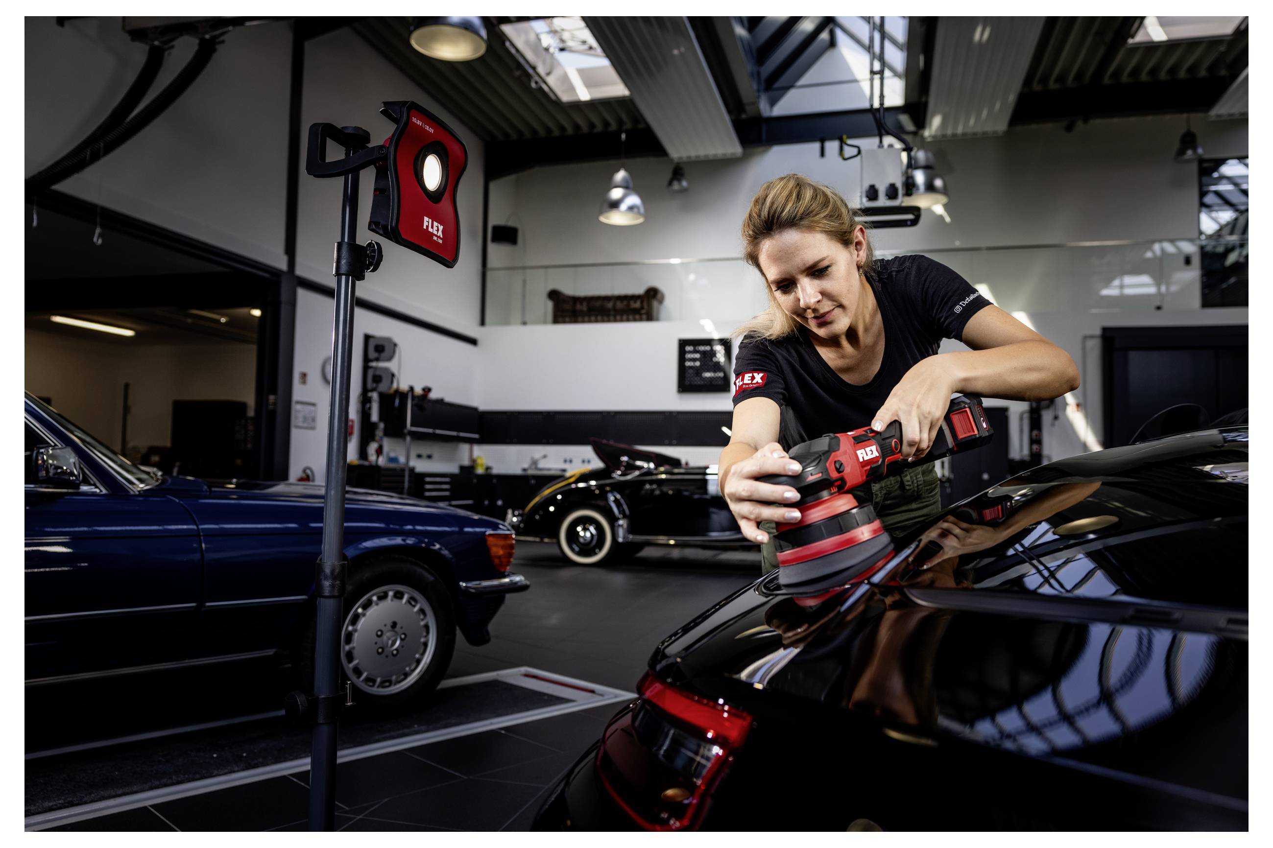 Žena leštící černé auto elektrickým leštidlem uvnitř garáže showroomu s dalšími klasickými auty v pozadí.