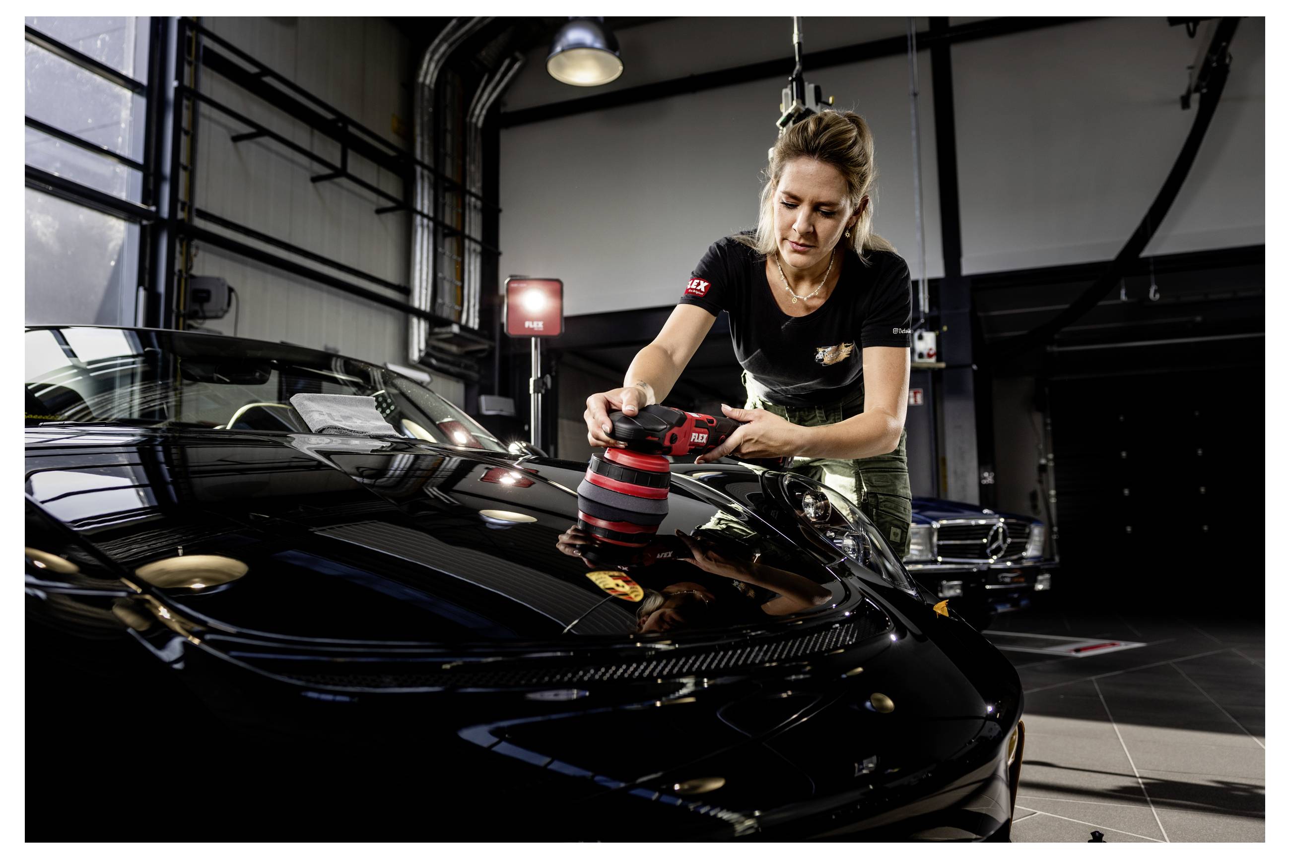 Žena používající ruční leštičku k broušení kapoty černého auta v autodetailingové dílně.