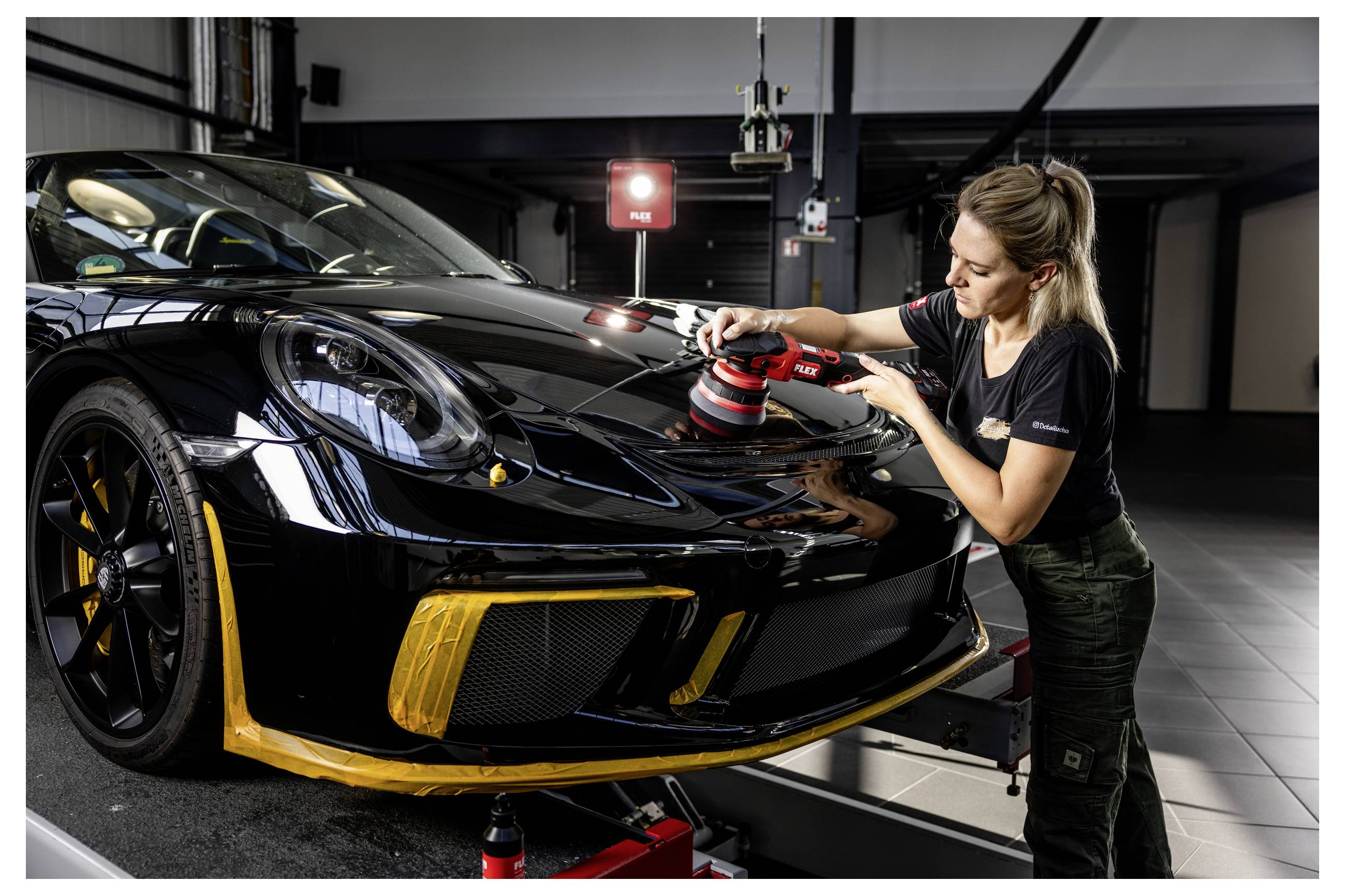 Žena leštící kapotu černého sportovního automobilu v garáži, používající červený leštící kotouč, zatímco je auto zvednuté na zvedáku.