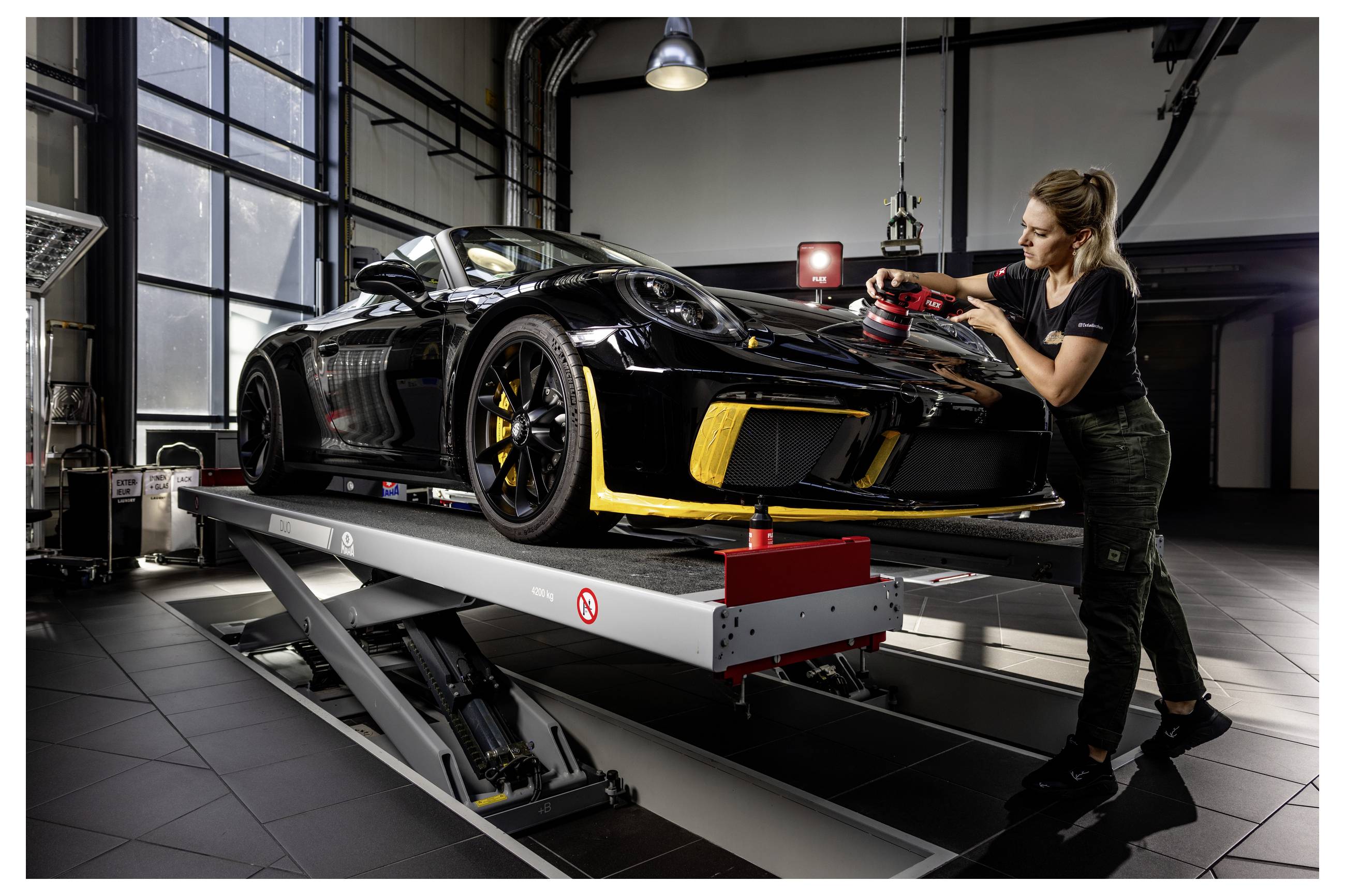 Technik leští přední část černého sportovního automobilu na hydraulickém zvedáku v moderní garáži nebo detailingovém servisu.