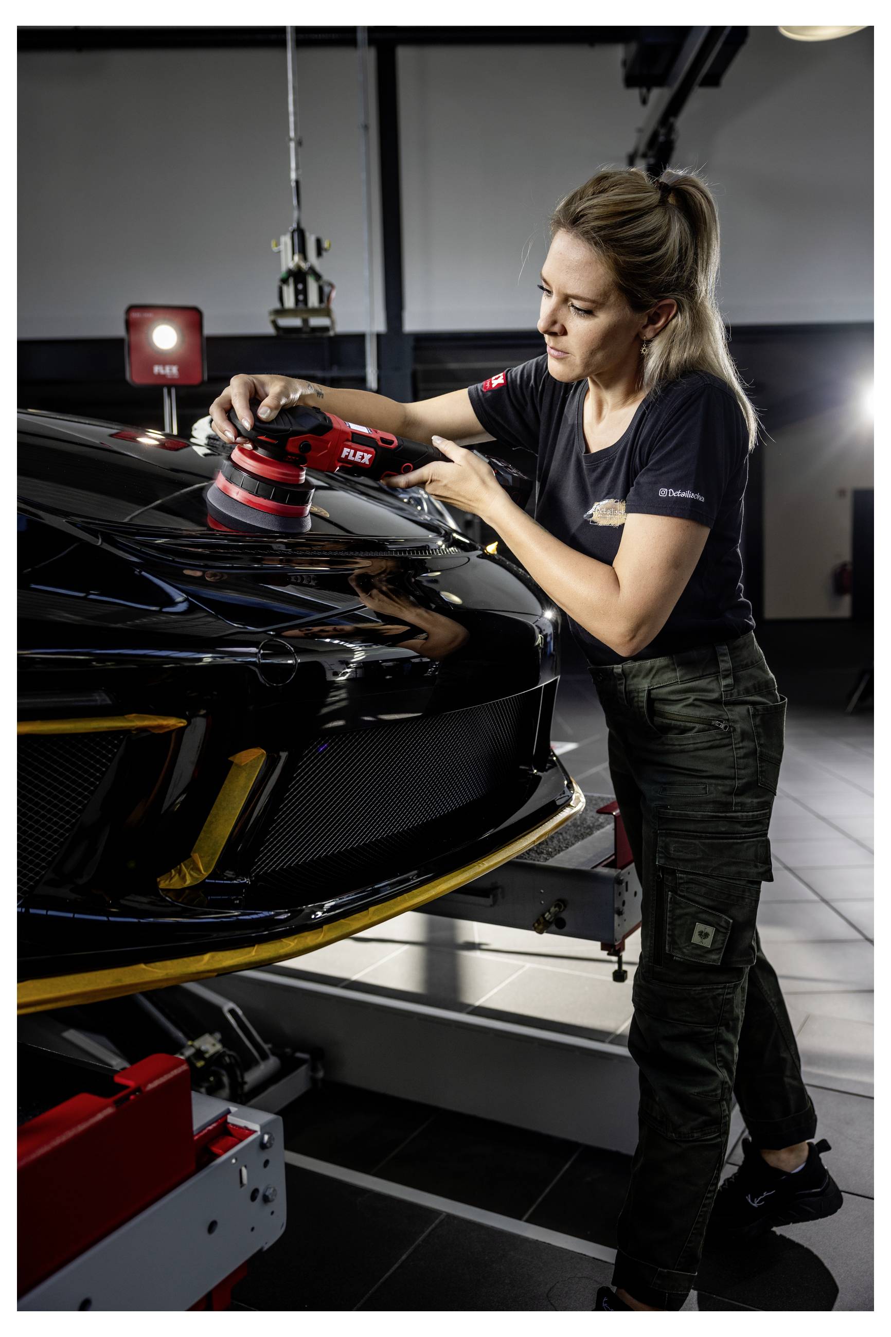 Žena leštící kapotu černého sportovního automobilu elektrickým leštičem v garáži nebo autopraní.