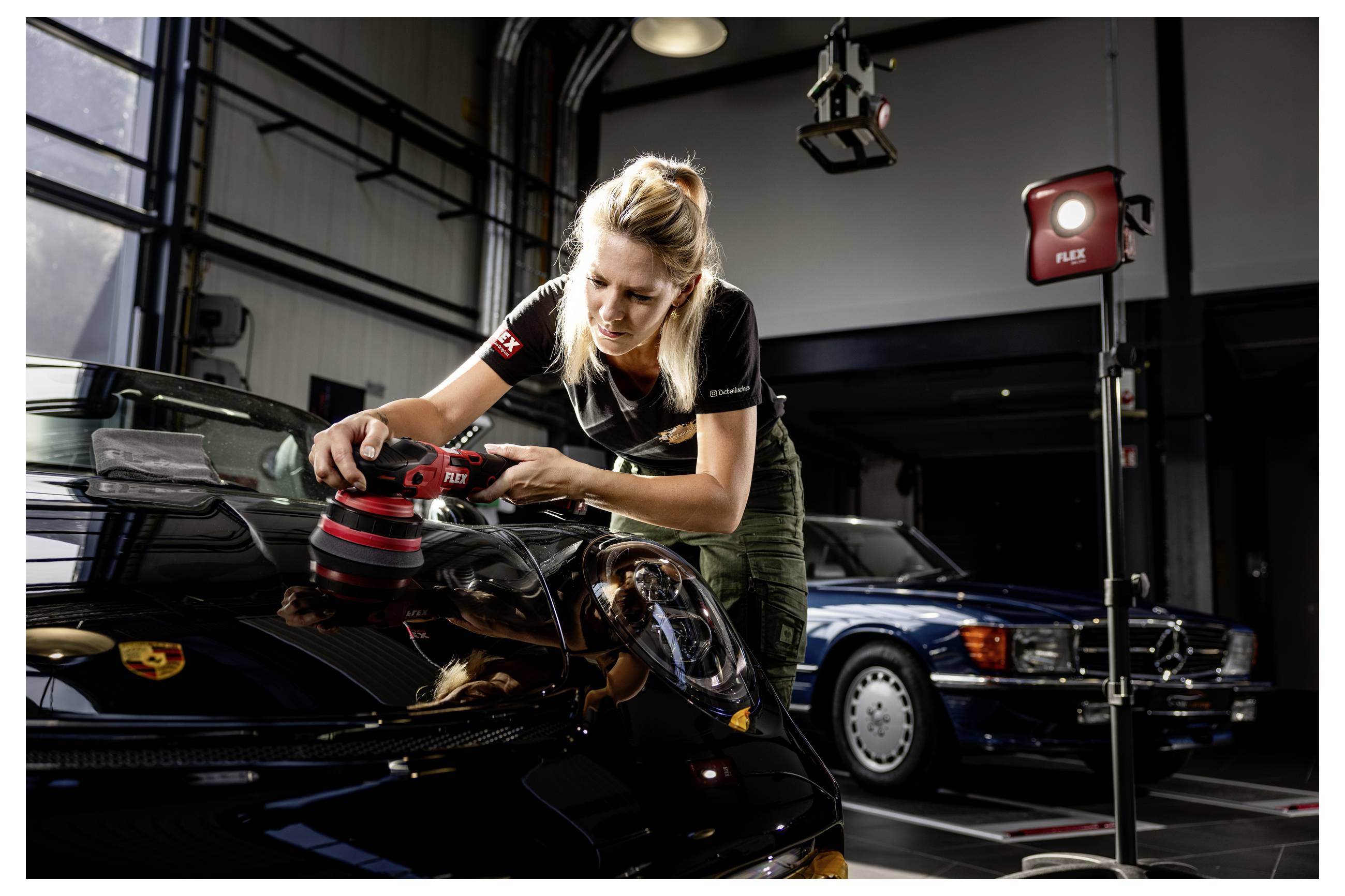 Žena leštící kapotu černého Porsche v dílně, která používá elektrickou leštičku vedle dalších zaparkovaných aut a studiových světel.