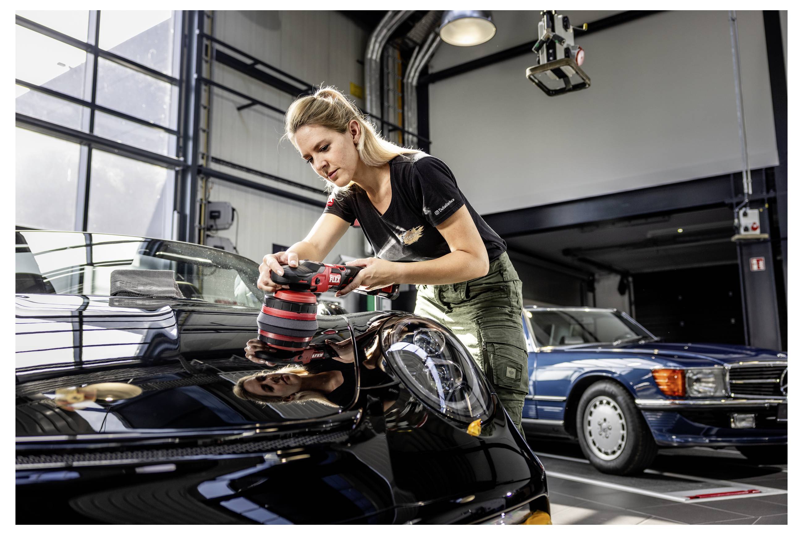 Žena leští kapotu černého automobilu v garáži, v pozadí je zaparkován další historický automobil.