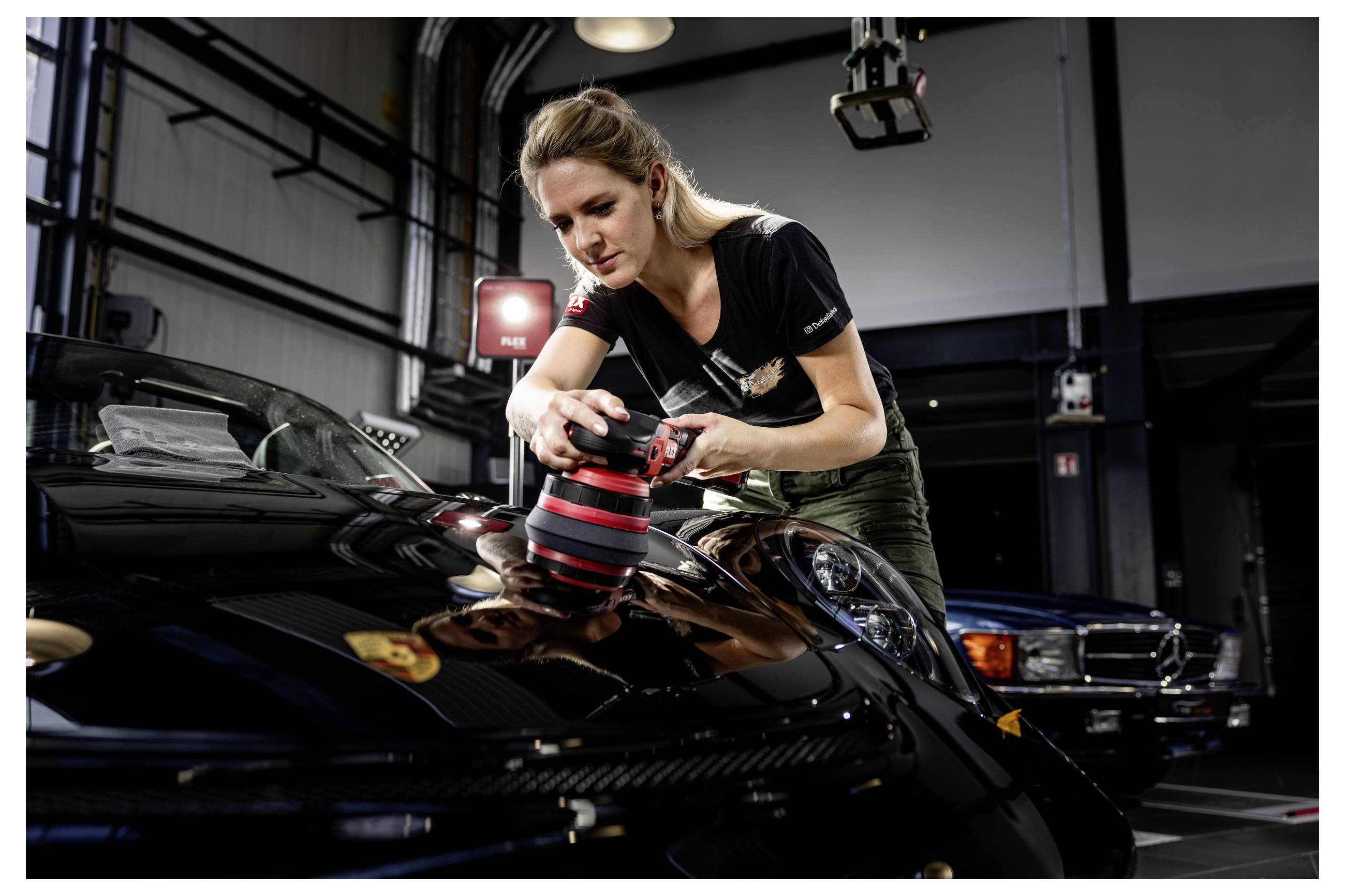Žena leštící černý sportovní vůz elektrickým leštidlem v autokempu se soustředí na detailní úpravu kapoty.