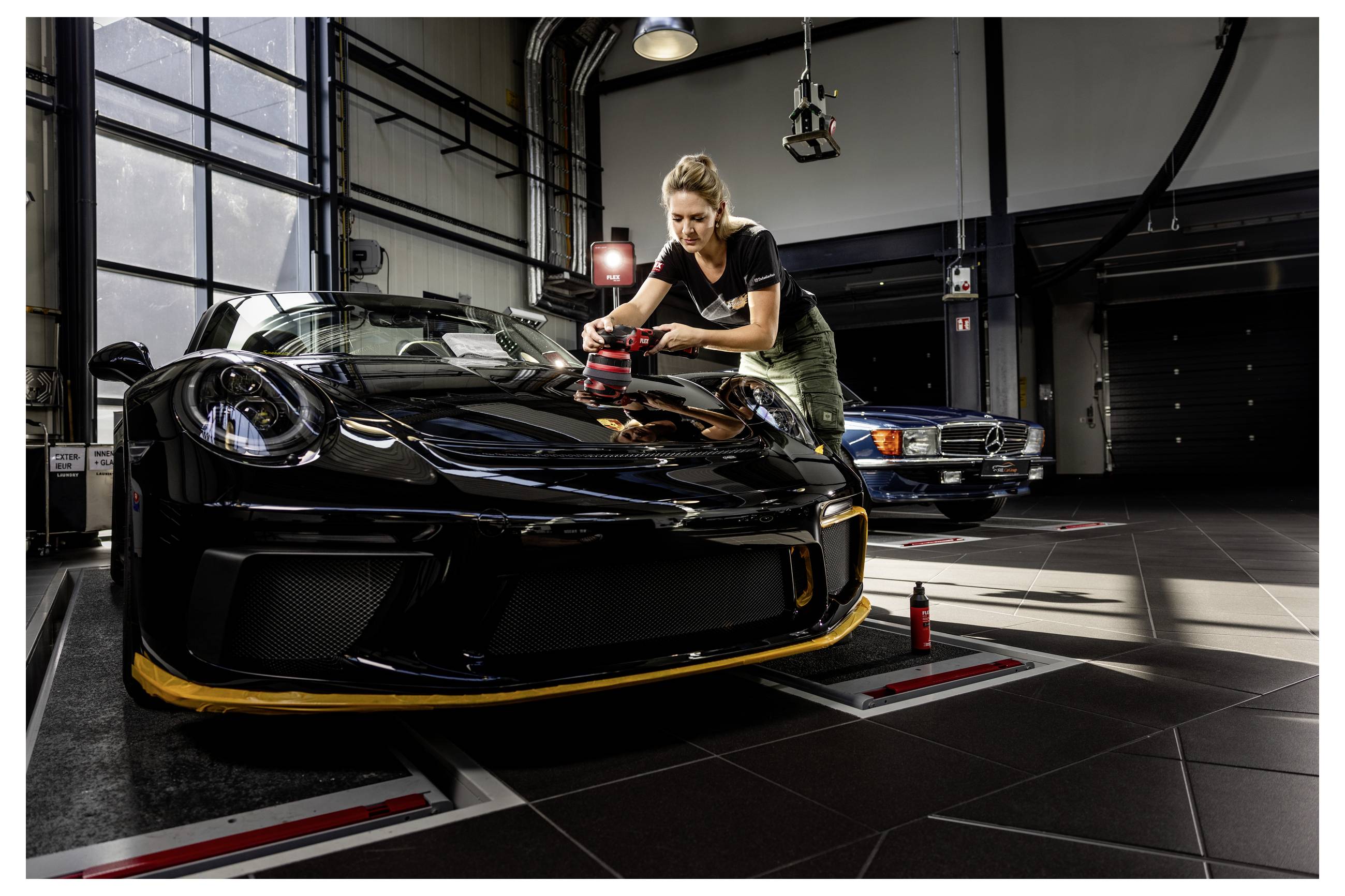 Žena leští kapotu černého sportovního auta v garáži, přičemž za ní je zaparkované další klasické auto.