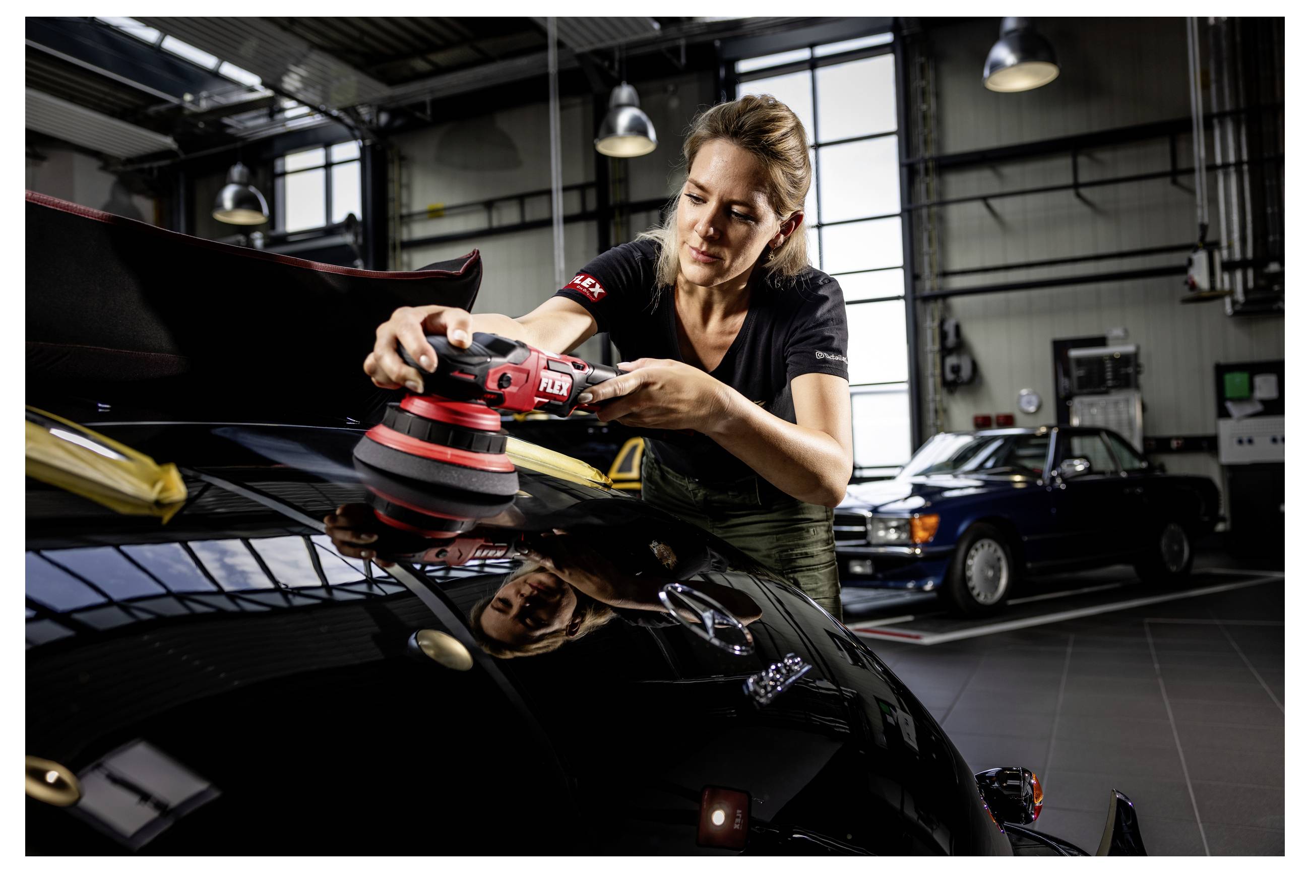 Žena leštící kapotu černého sportovního automobilu elektrickým leštidlem v garáži, s dalším autem v pozadí.