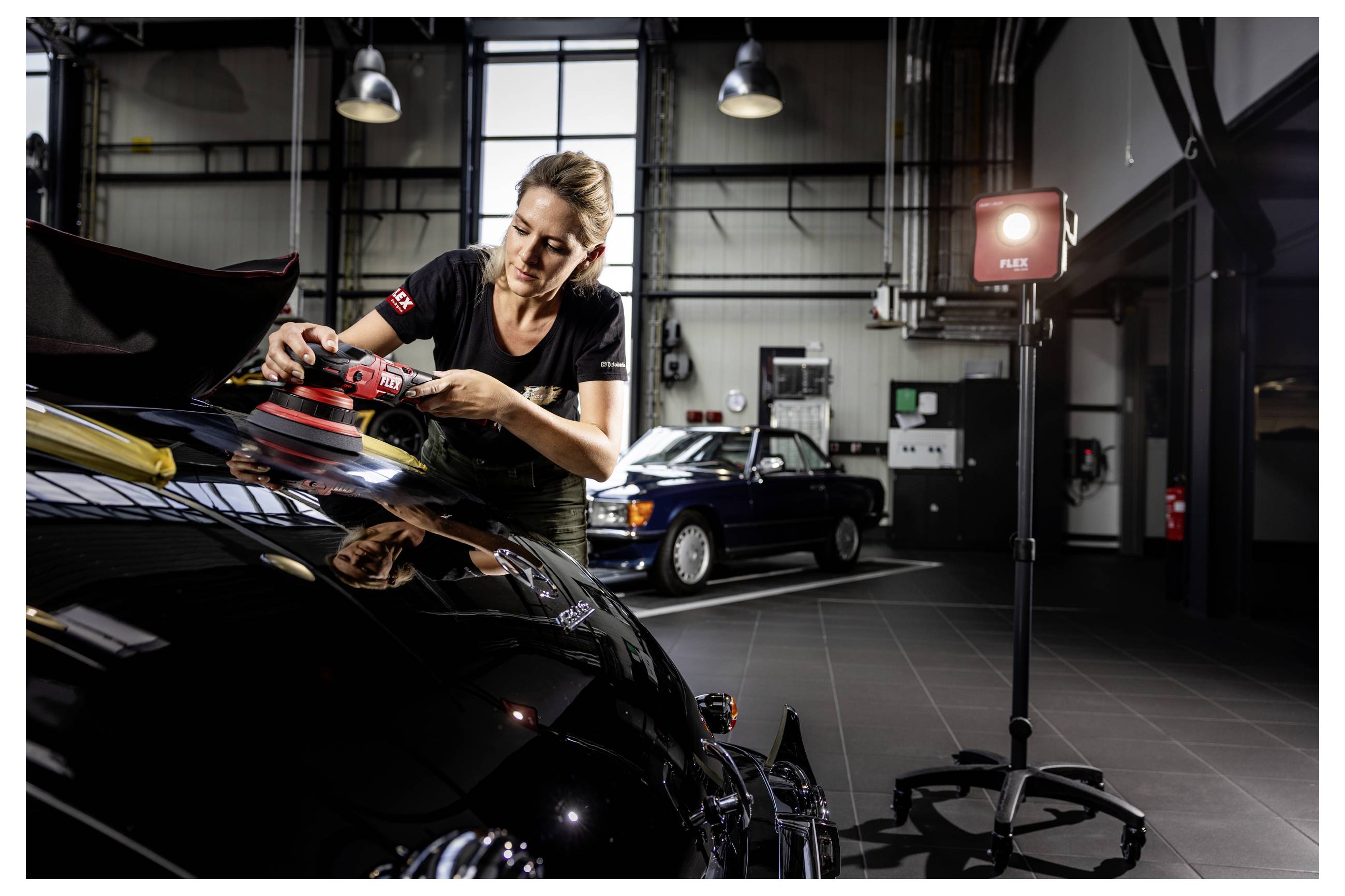 Automechanik vyleštuje černé auto elektrickým leštidlem v autoservisu, v pozadí jsou další vozidlo a tabule s nápisem "FIRE".