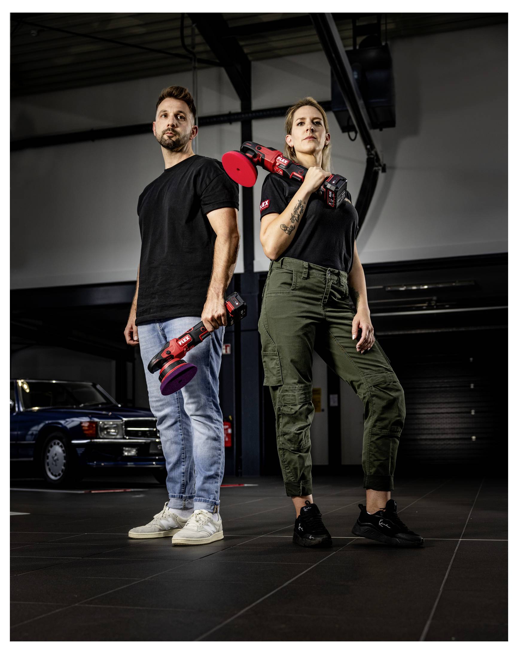 Dva mechanici stojí v garáži a drží červené elektrické leštičky, postaveni zády k sobě vedle klasického automobilu.