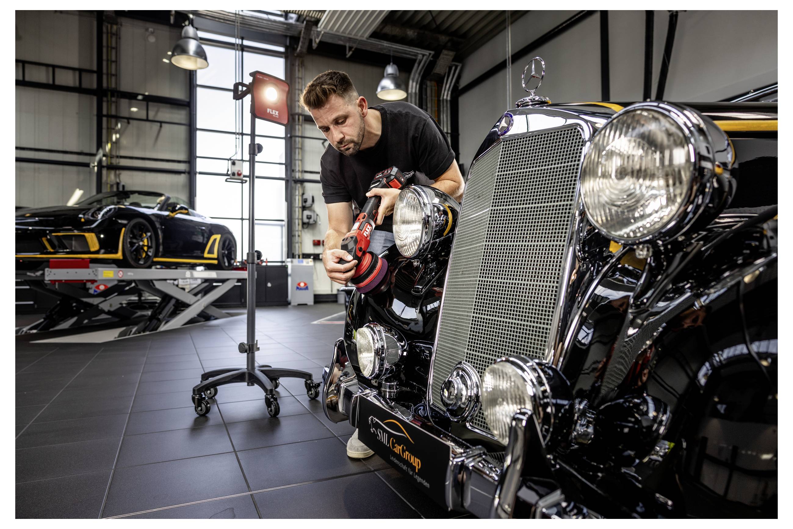Mechanik leštící přední část klasického černého automobilu v garáži, s dalším sportovním vozem na zvedáku v pozadí.