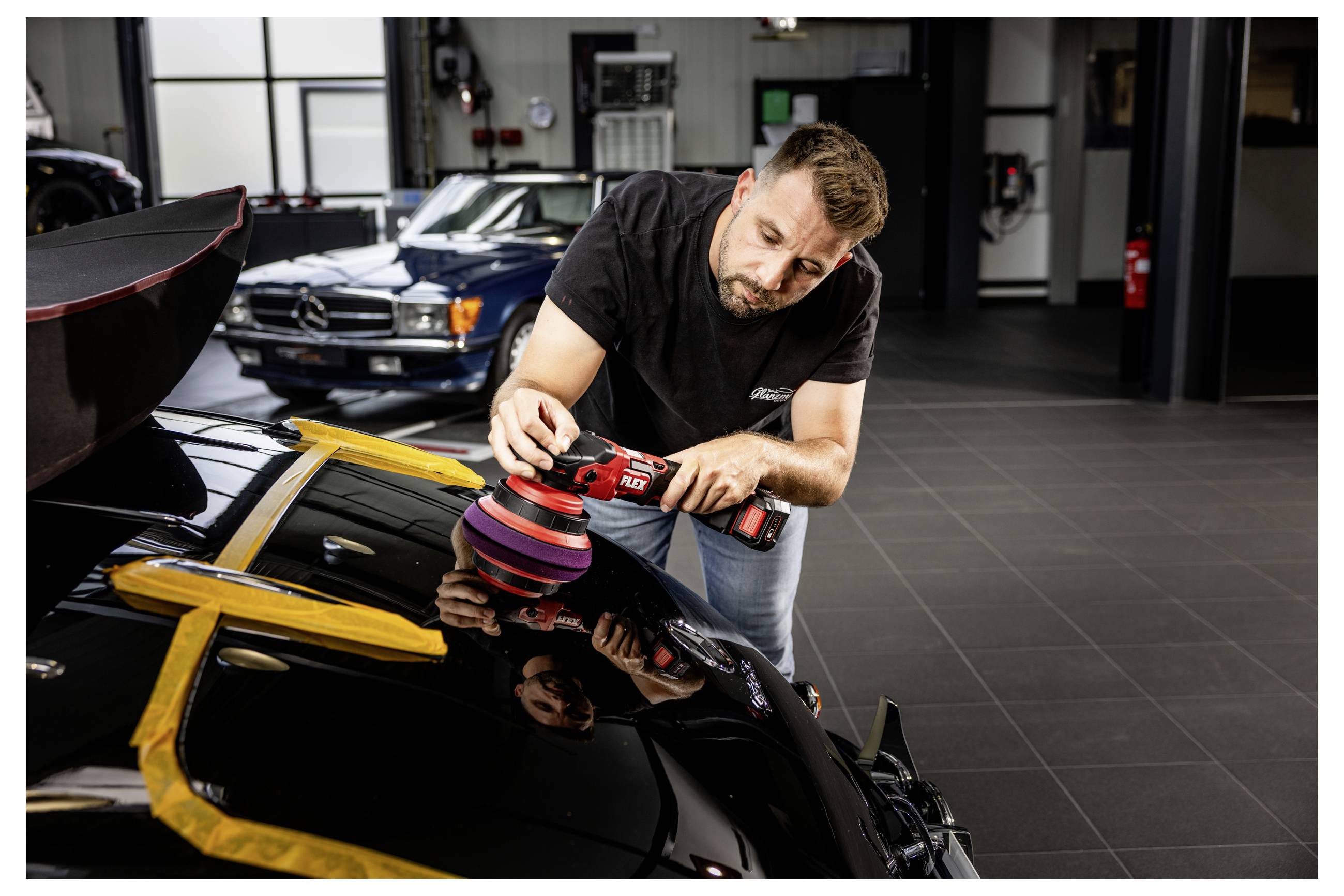 Muž leští černý sportovní vůz elektrickým leštidlem v garáži, v pozadí je vidět další klasický automobil.