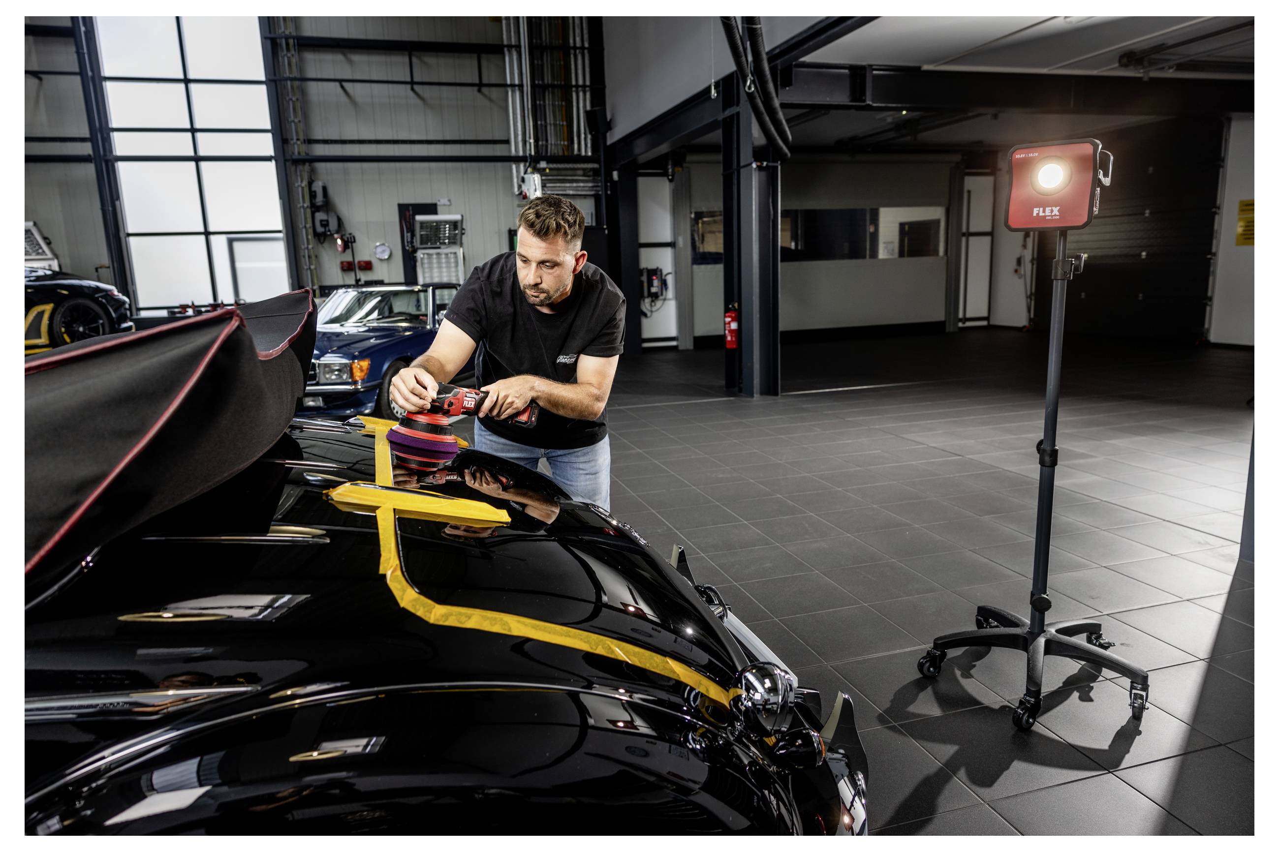 Muž leští kapotu černého automobilu s leštícím nástrojem v garáži, poblíž se nachází červené pracovní světlo na stojanu.