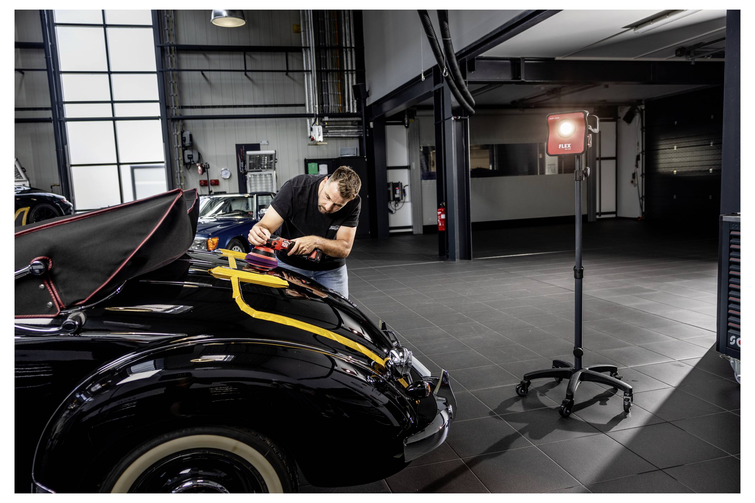 Automechanik leští černý automobil v dílně, přenosné inspekční světlo osvětluje vozidlo.