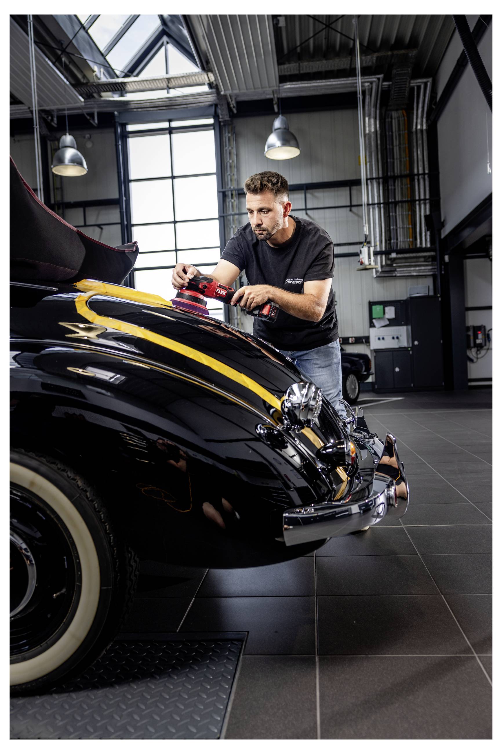 Muž leštící černý vintage automobil se žlutými proužky v garáži podobné dílně.