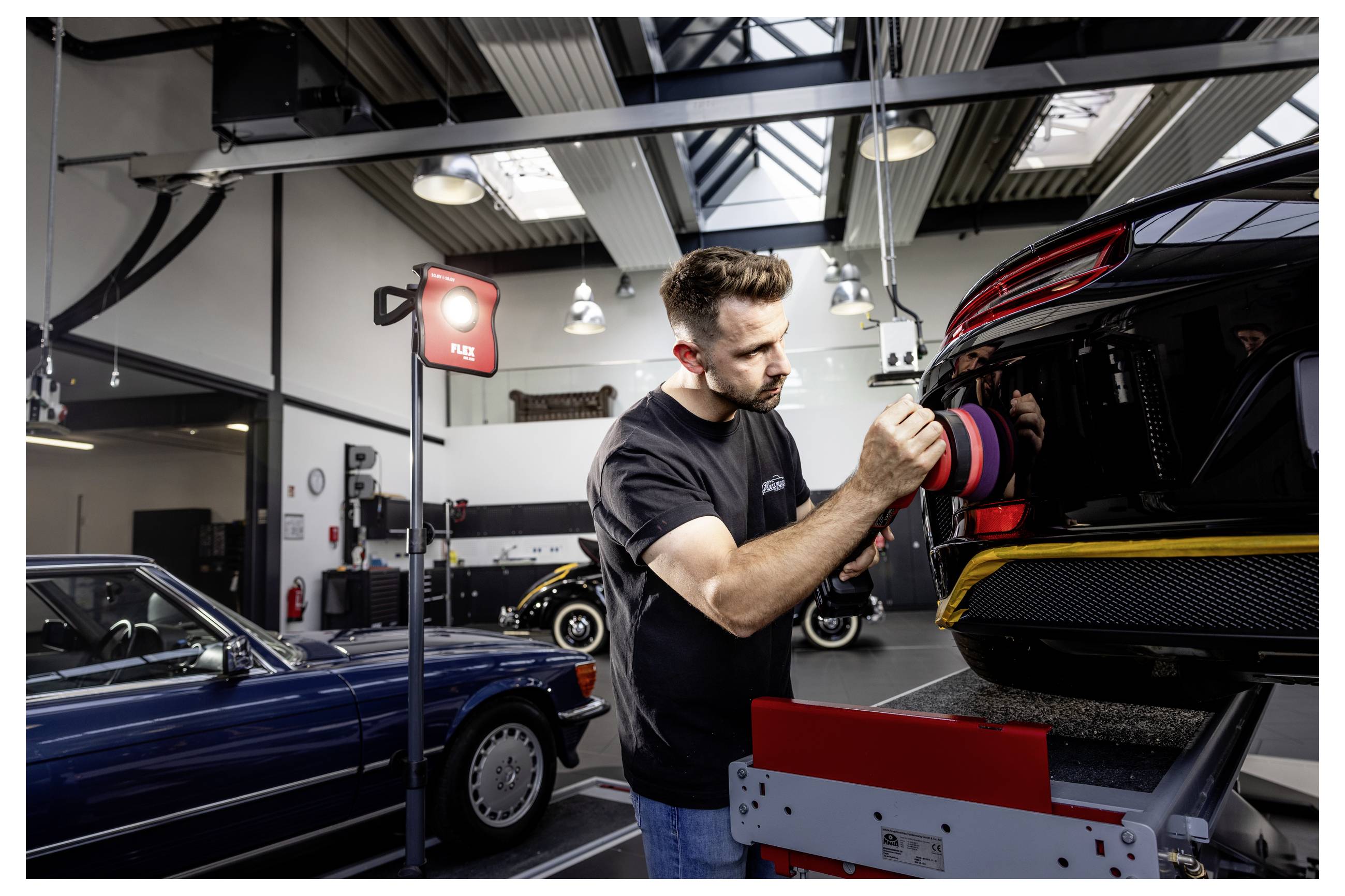 Automechanik leští zadní část černého sportovního automobilu pomocí leštičky uvnitř autodílny, v pozadí jsou další automobily.