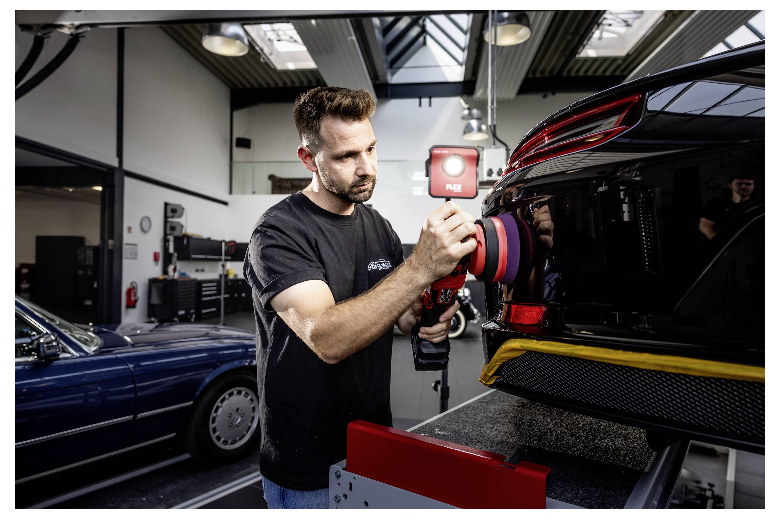 Automechanik leštící černé auto elektrickým bufferem v dílně, s dalším vozidlem zaparkovaným v pozadí.