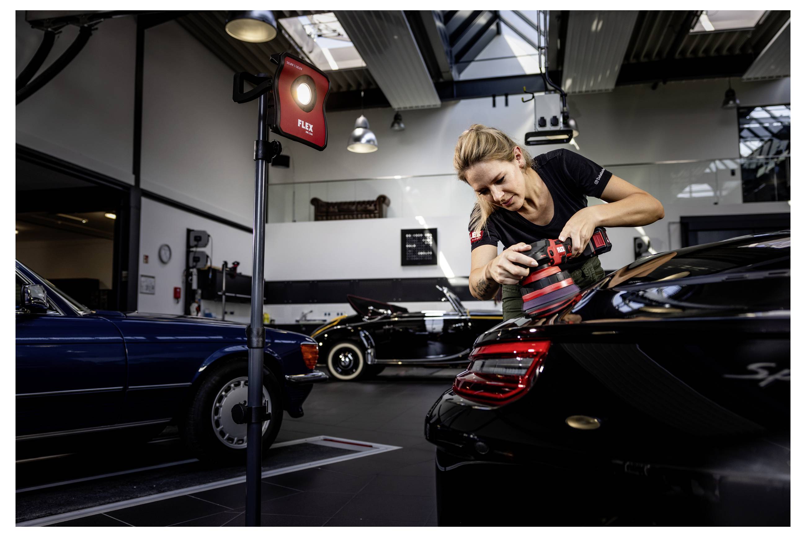 Žena leštící černý sportovní automobil s leštičkou uvnitř autodílny na detailní čištění aut, s dalšími klasickými auty v pozadí.