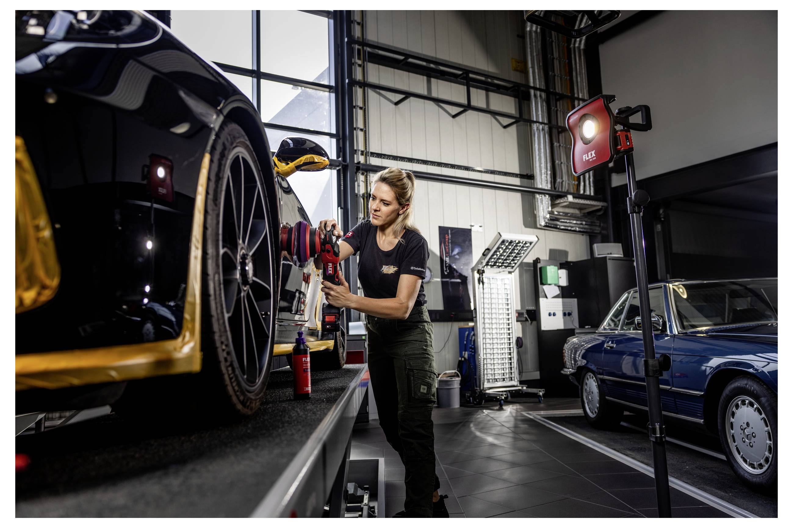 Automechanik upravuje přední kolo a brzdový systém černého sportovního vozu v garáži s dalším klasickým autem a dílenskými světly v pozadí.