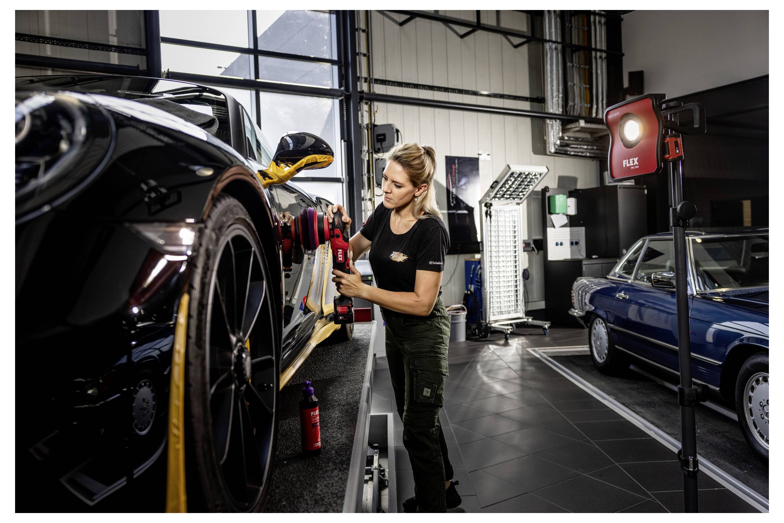 Mechanik leštující černý sporťák v autopracoviště, s dalším klasickým autem a pracovními světly v pozadí.