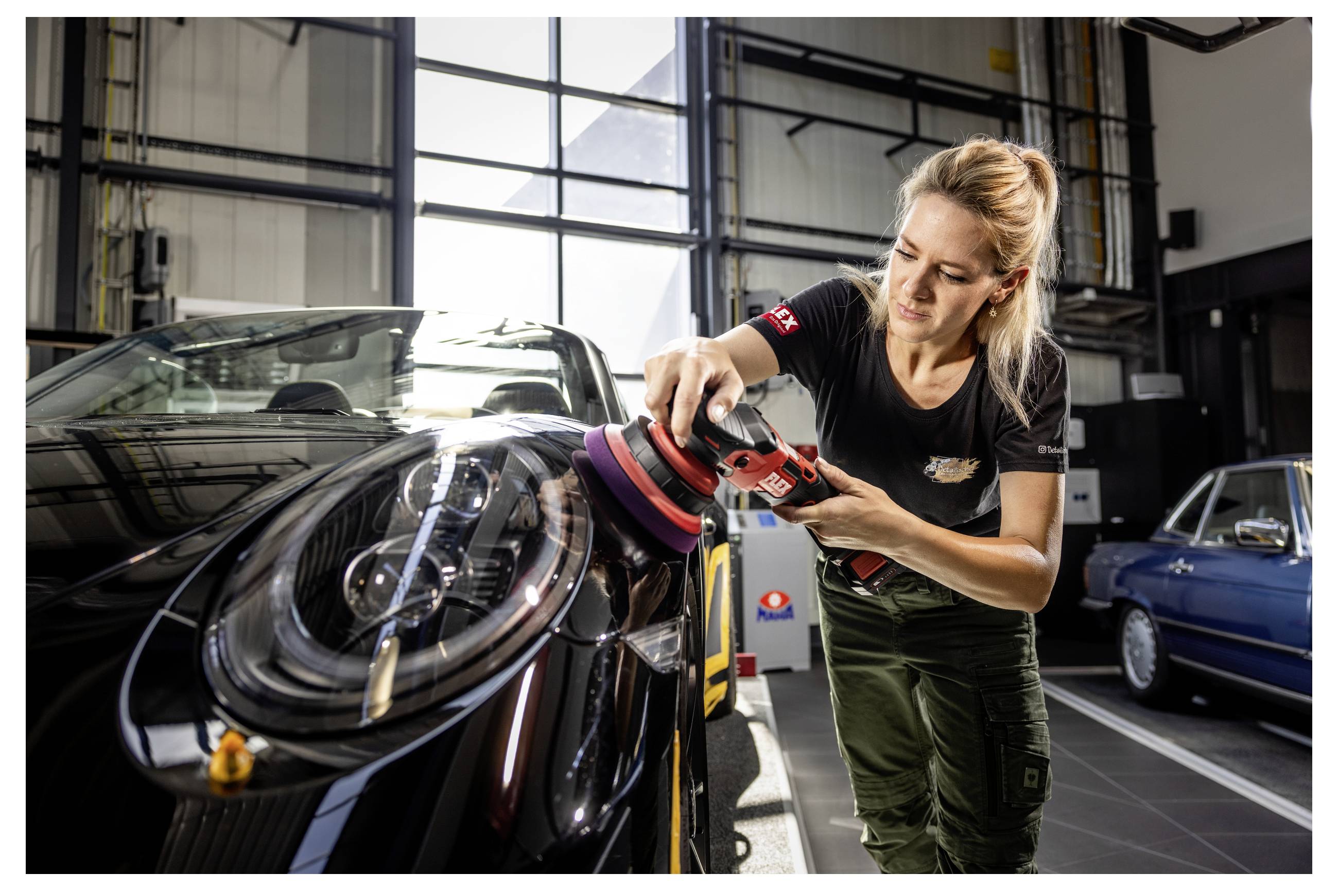 Žena leští přední část černého sportovního vozu elektrickou leštičkou uvnitř autoservisu.