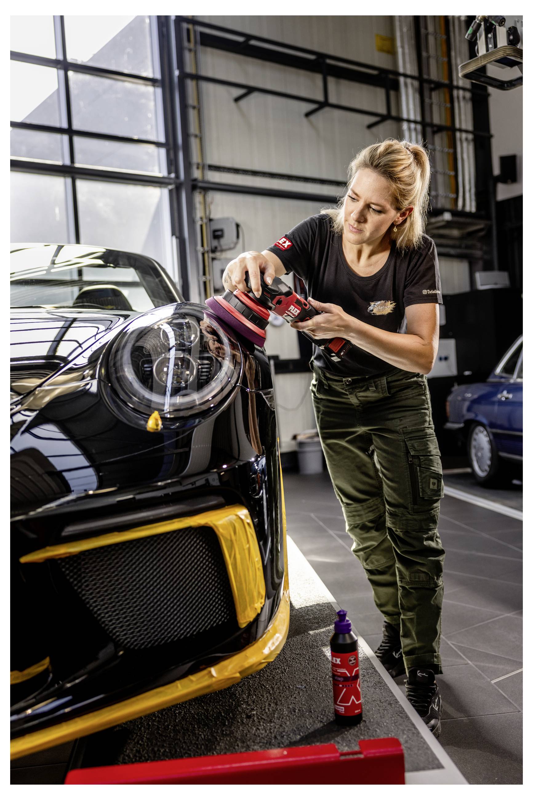 Žena leštící přední část černého sportovního automobilu v garáži, používající červeno-černý elektrickýnářadí, s detailingovými pomůckami v blízkosti.