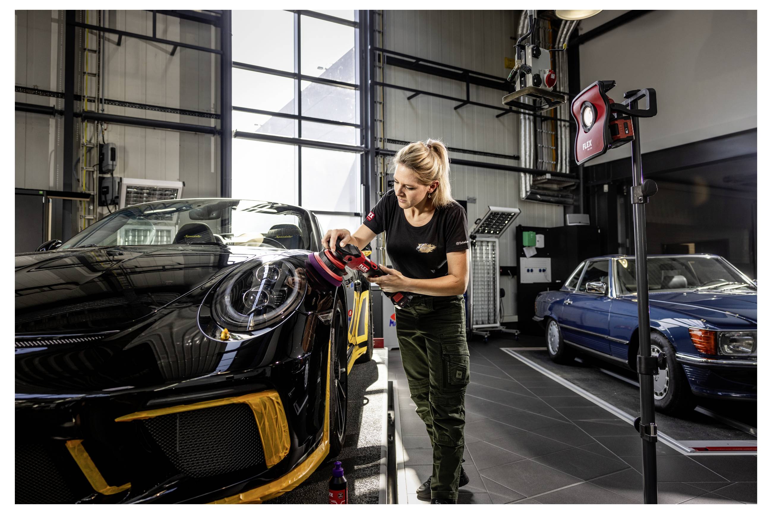 Žena leští černé sportovní auto v garáži, poblíž je zaparkované další klasické modré auto.