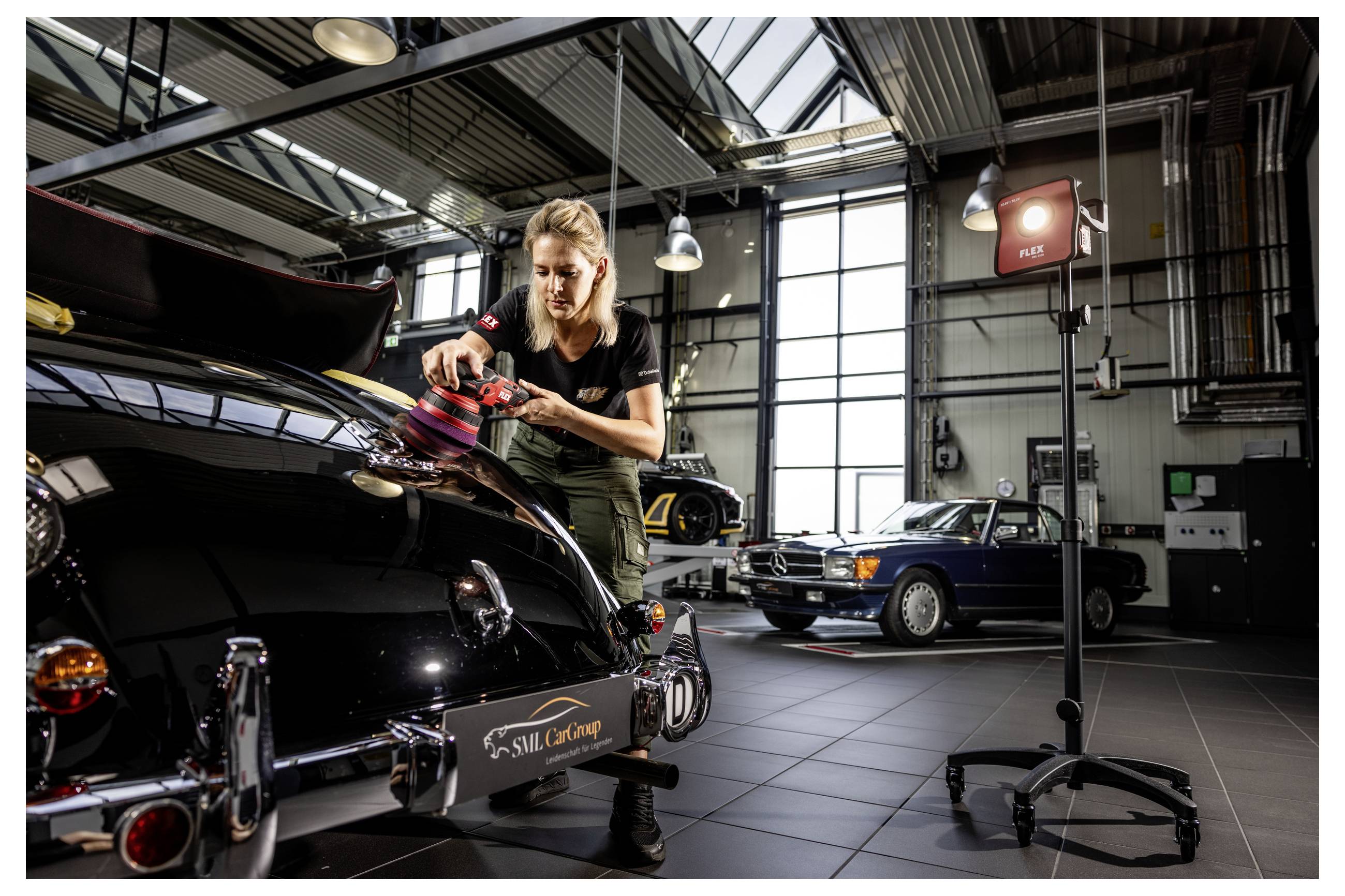 Mechanik leští kapotu černého klasického vozu v jasné automechanické dílně s dalším vintage autem v pozadí.