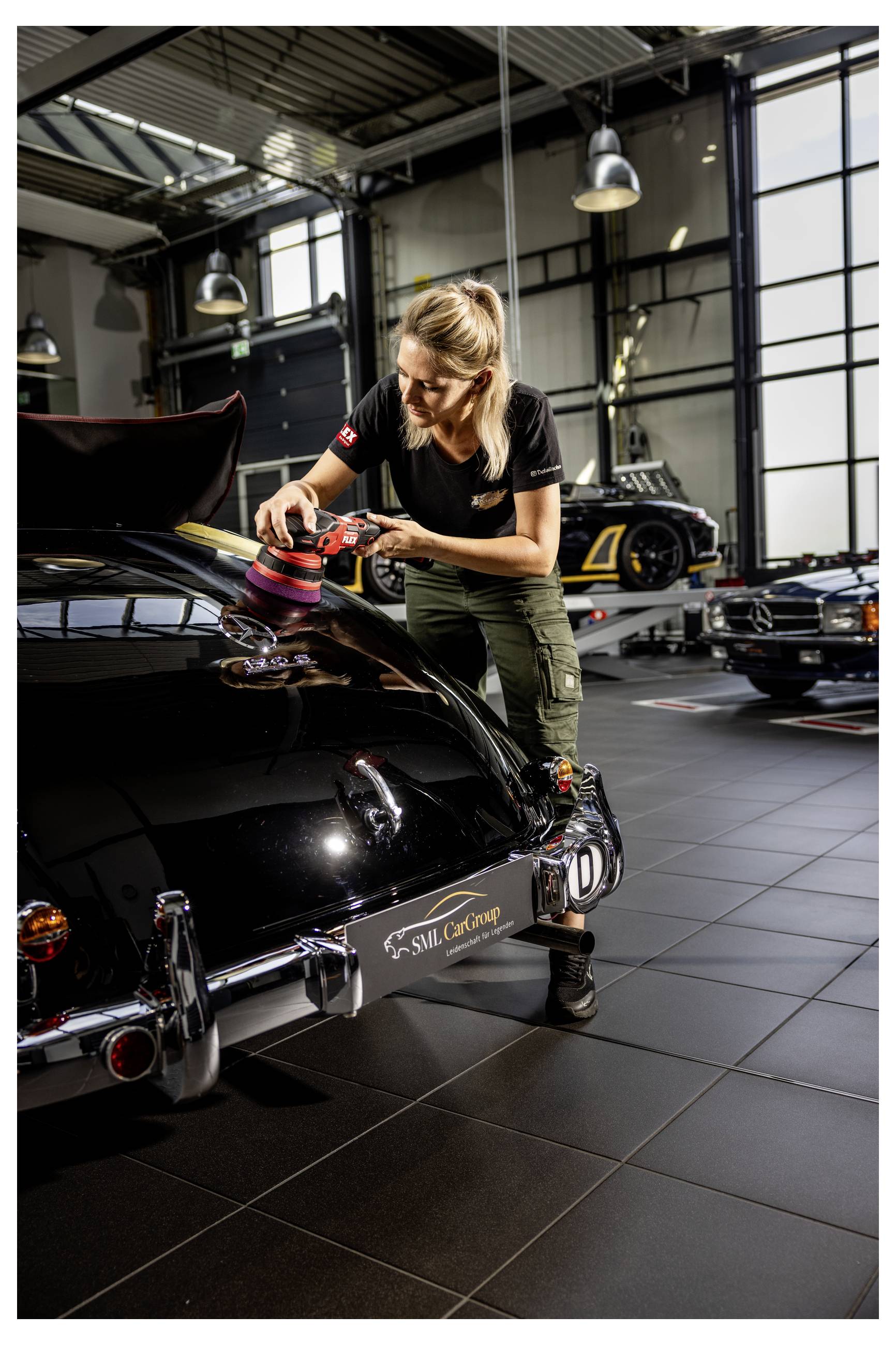 Žena leštící zadní část černého veteránského vozu v garáži, s dalšími klasickými automobily a pracovními světly v pozadí.