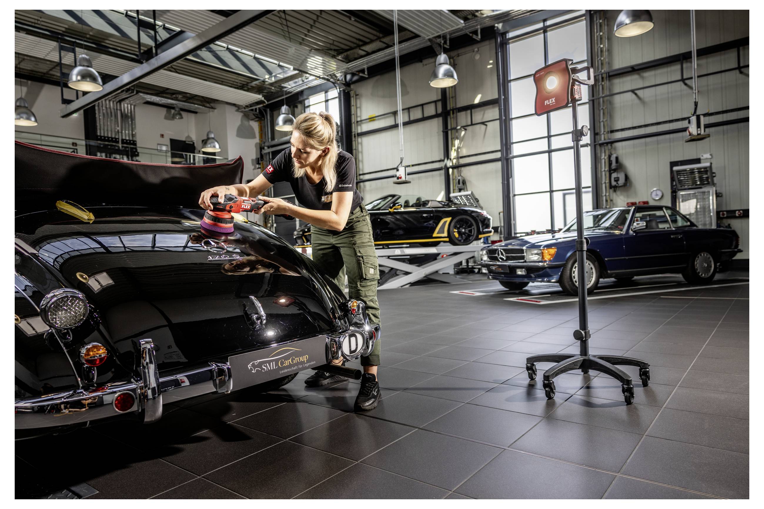Žena leštící černý klasický automobil s leštičkou v dobře osvětleném autosalónu, s dalšími veterány v pozadí.