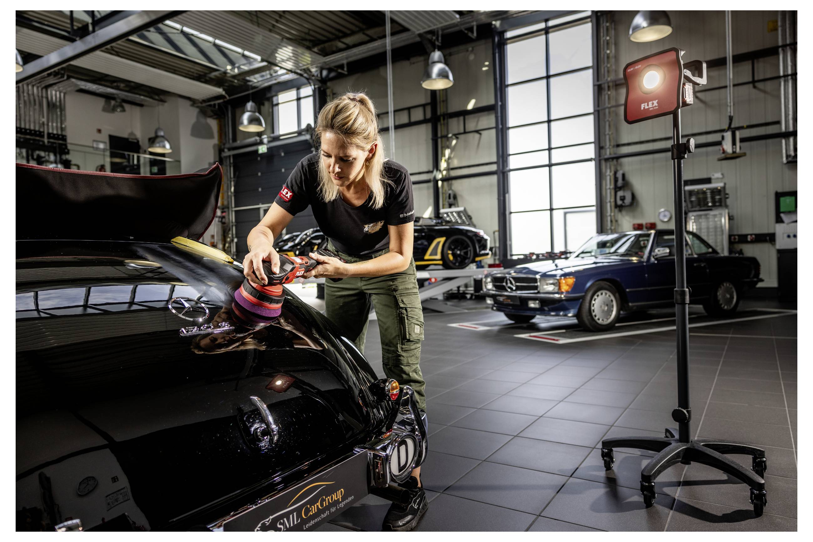 Žena leštící kapotu černého automobilu v garáži dílny s dalšími klasickými automobily zaparkovanými v pozadí.
