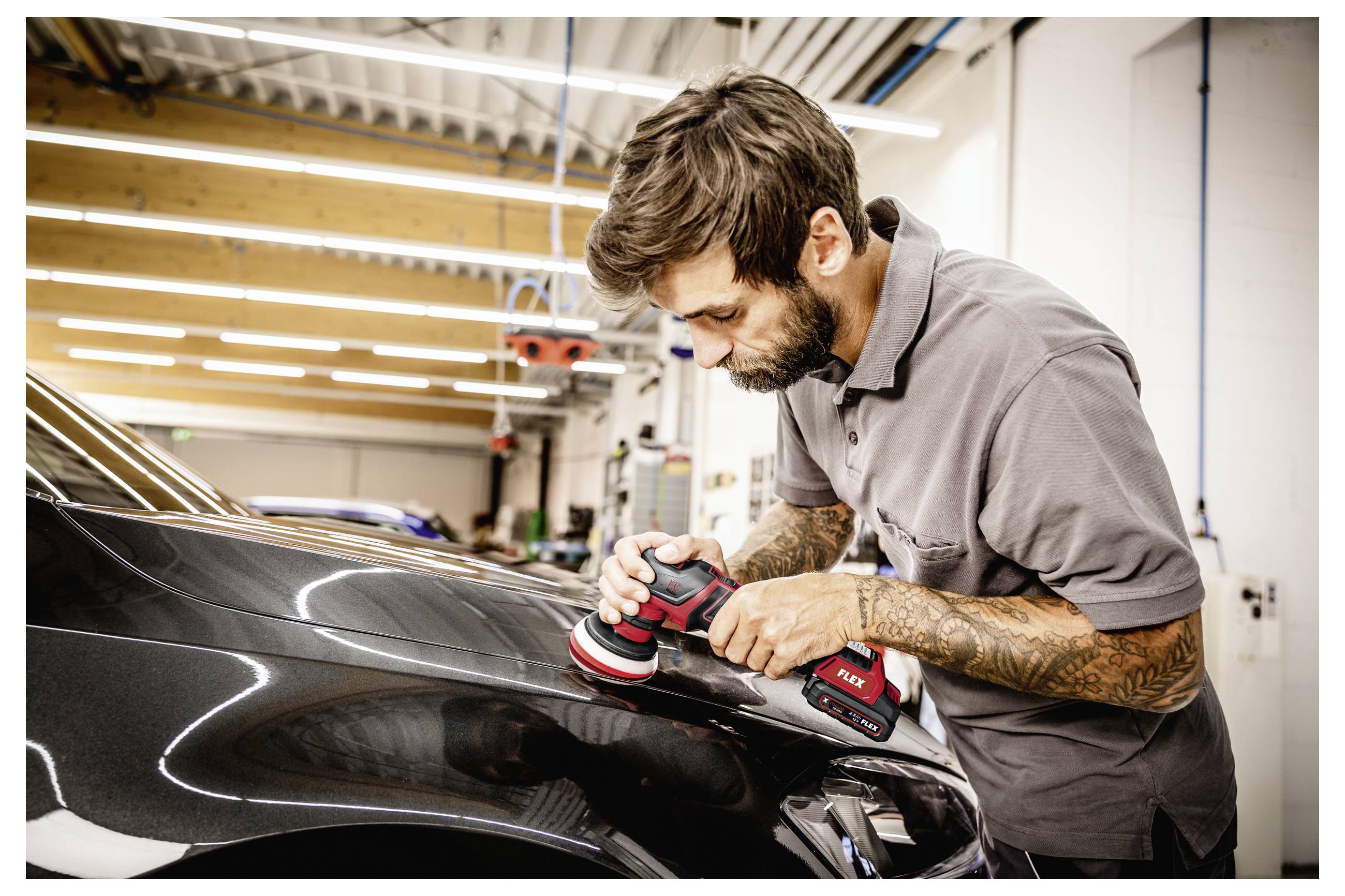Muž leští kapotu černého automobilu ruční leštičkou v autoopravně.
