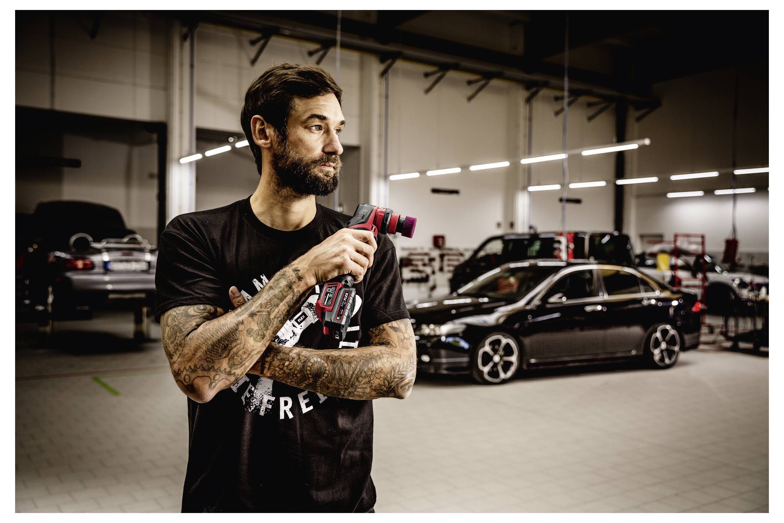 Tetovaný muž držící červený elektrický nástroj v autoservisu s několika černými auty zaparkovanými za ním.
