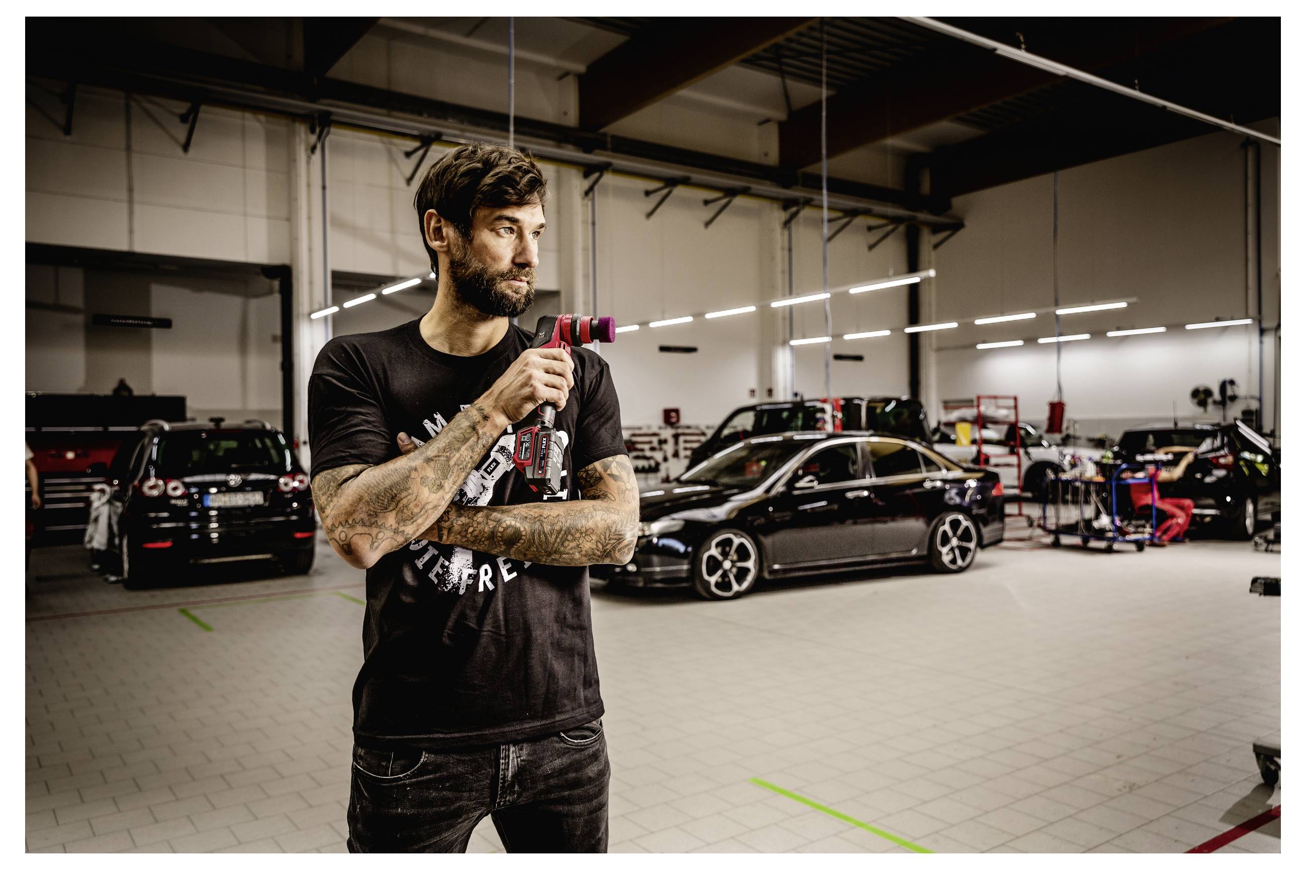 Tetovaný muž v černém tričku stojí v autoservisu a drží v ruce elektrickém nářadí, v pozadí jsou upravená auta a vybavení.