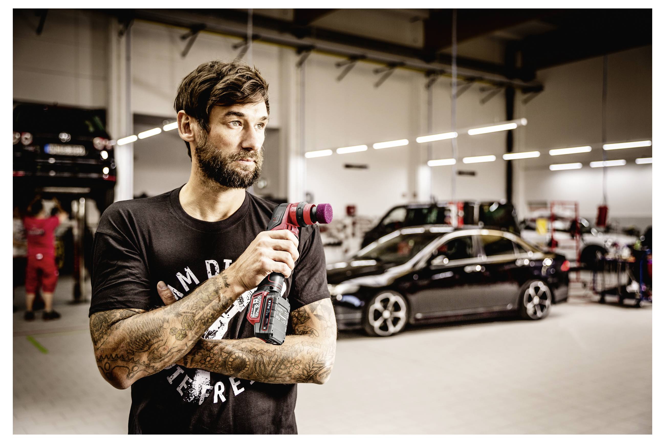 Tetovaný mechanik držící bezšňůrový nástroj v autoservisu s vozidly a vybavením v pozadí.
