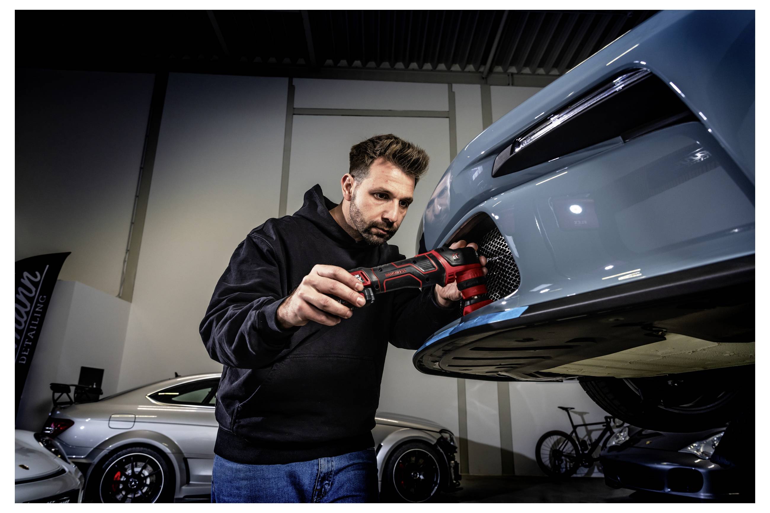 Mechanik používá elektrické nářadí na přední části modrého sportovního vozu v garáži, v pozadí jsou viditelná další vozidla.