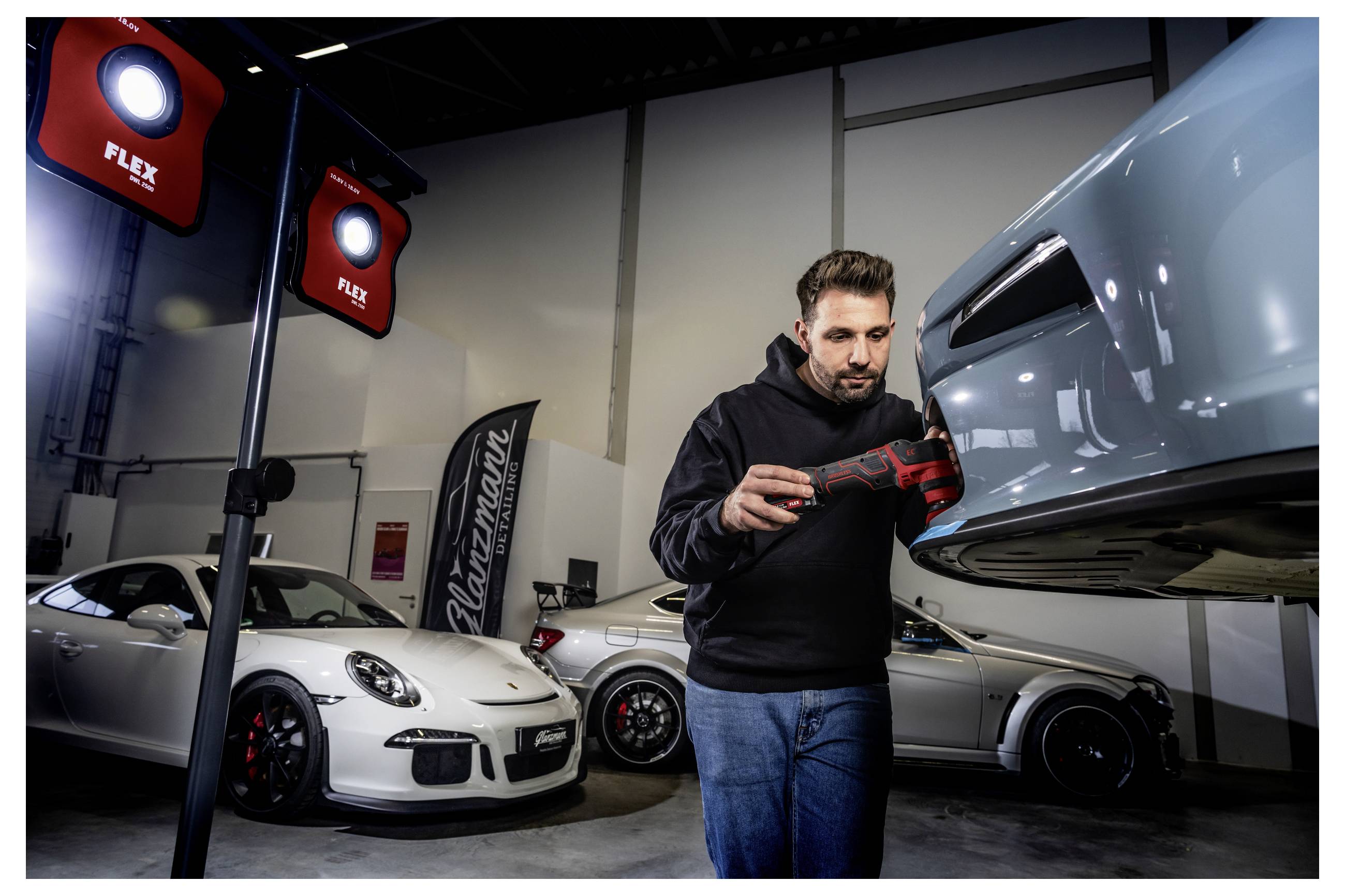 Muž kontroluje a leští přední část světle modré sportovního automobilu v garáži se dvěma dalšími auty Porsche a červenými pracovními svítidly FLEX v pozadí.