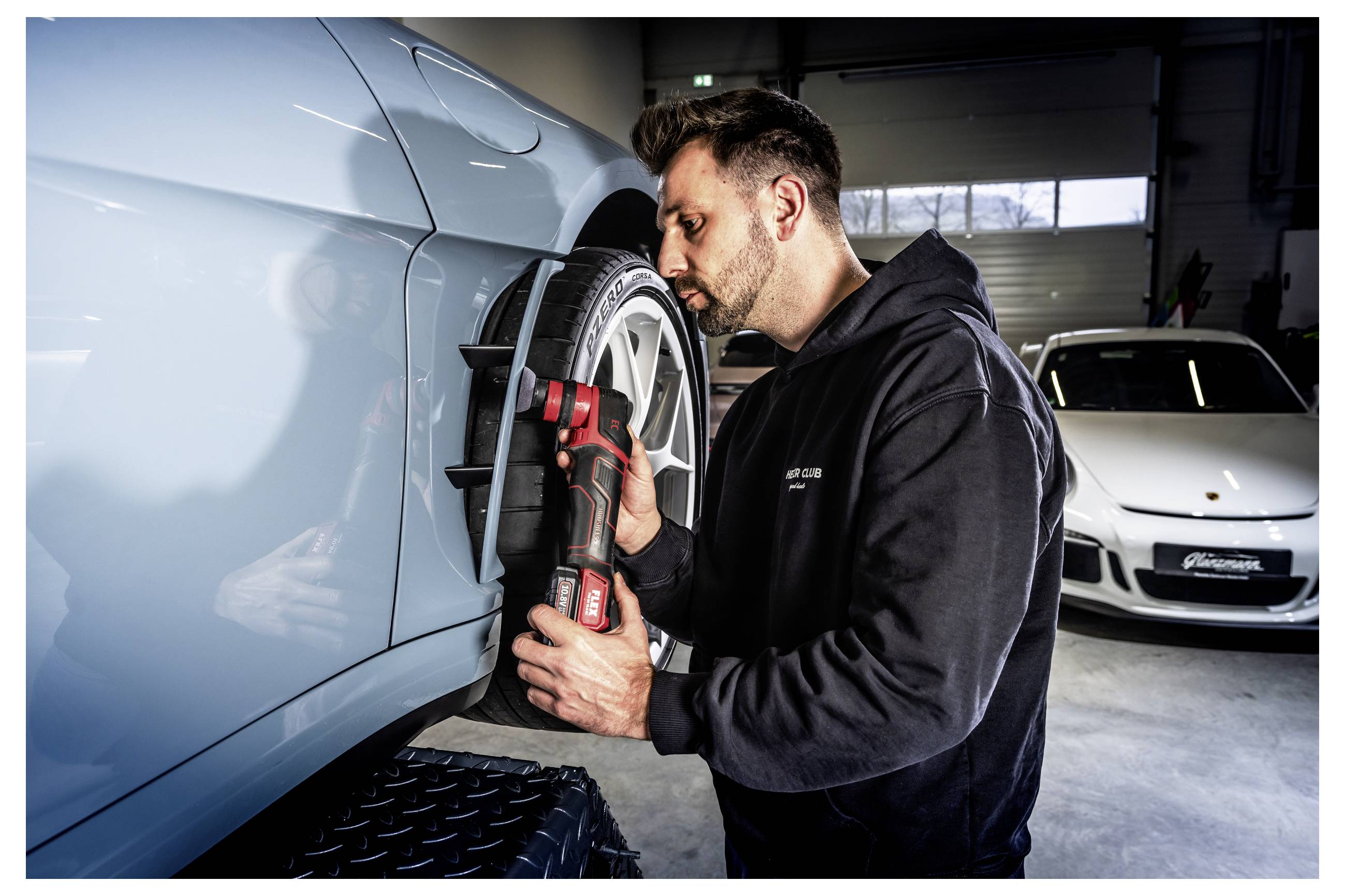 Mechanik kontroluje a leští oblast kol světlemodrého auta v garáži, v pozadí je zaparkované bílé sportovní auto.