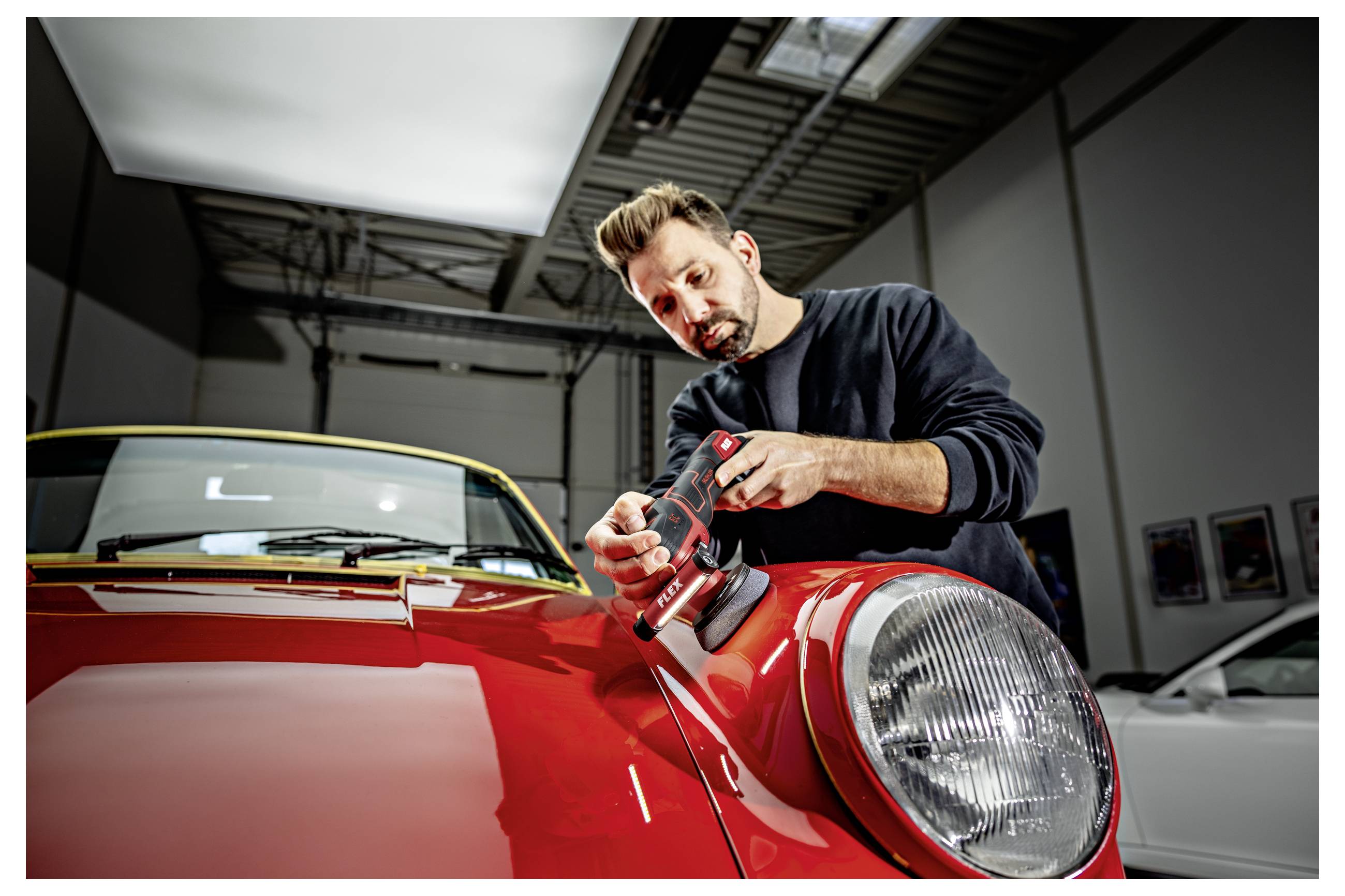 Automechanik leštící kapotu červeného klasického automobilu v garáži, používající přenosný leštič poblíž předního světlometu.