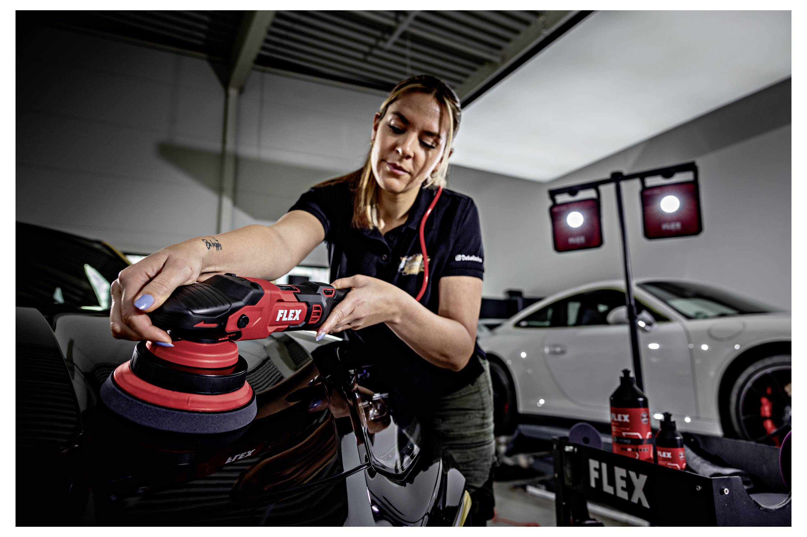 Technik používající FLEX leštičku k vyleštění auta v garáži, s bílým sportovním vozem a detailingovými potřebami v pozadí.