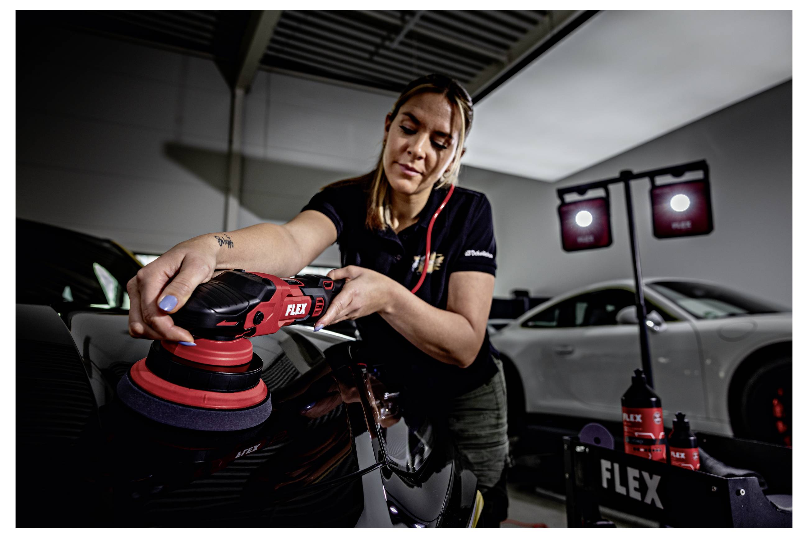 Žena leštící černé auto s červeným FLEX leštičem v garáži, s detailingovými potřebami a bílým autem v pozadí.