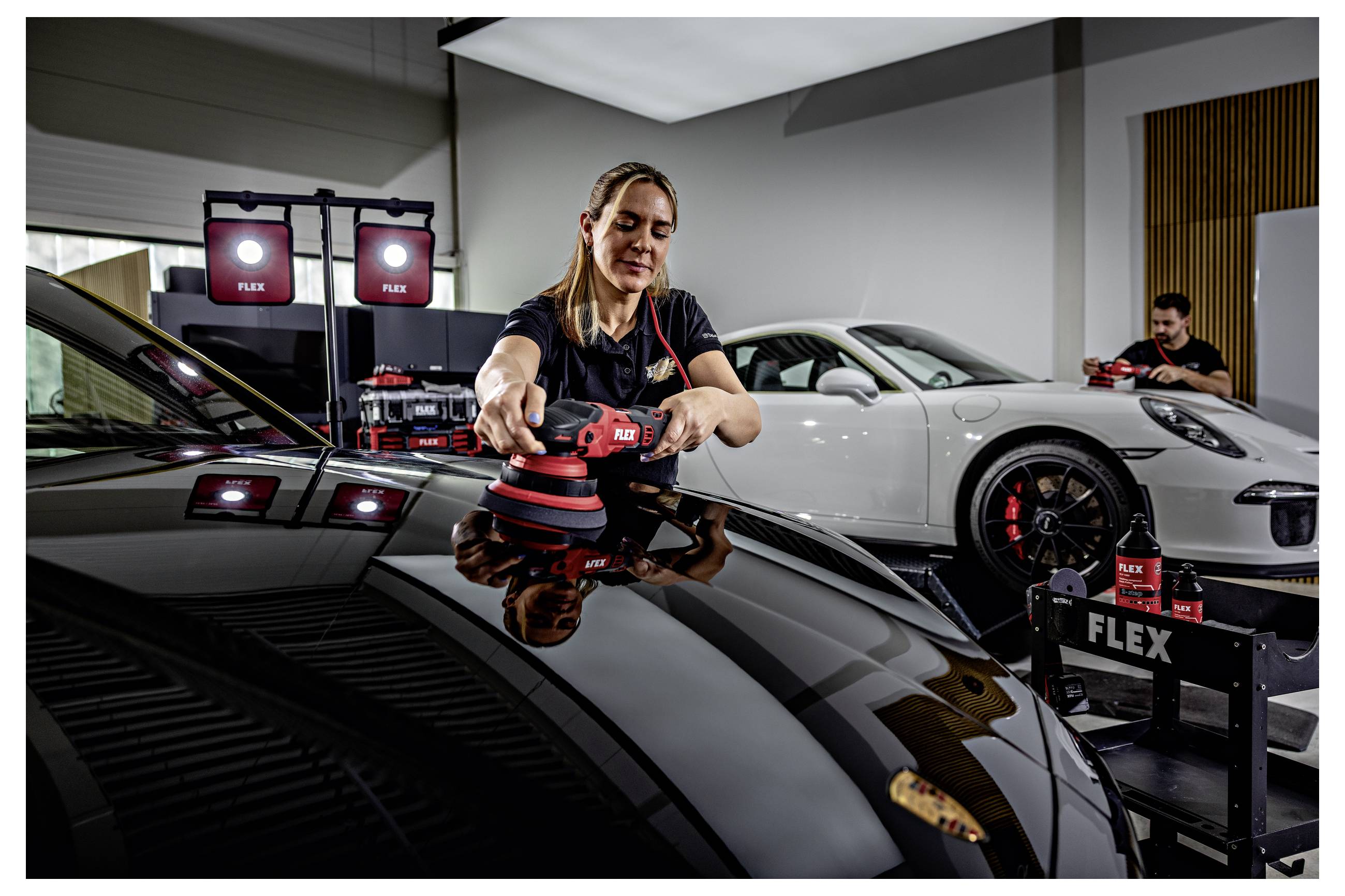 Technik leštící černý Porsche elektrickým leštičem FLEX v detailingové dílně, s bílým Porsche zaparkovaným za ní.