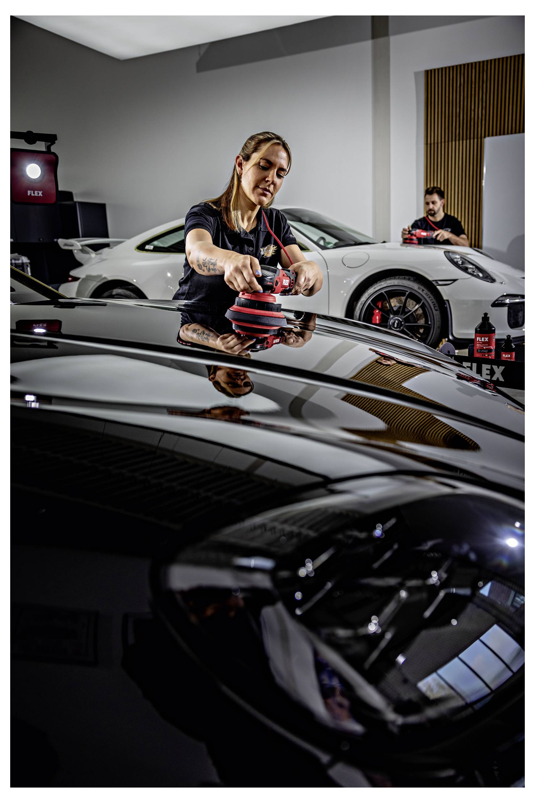 Technik detailingu leští černou kapotu auta elektrickým leštidlem v dílně, v pozadí je bílý sportovní vůz a další pracovník.