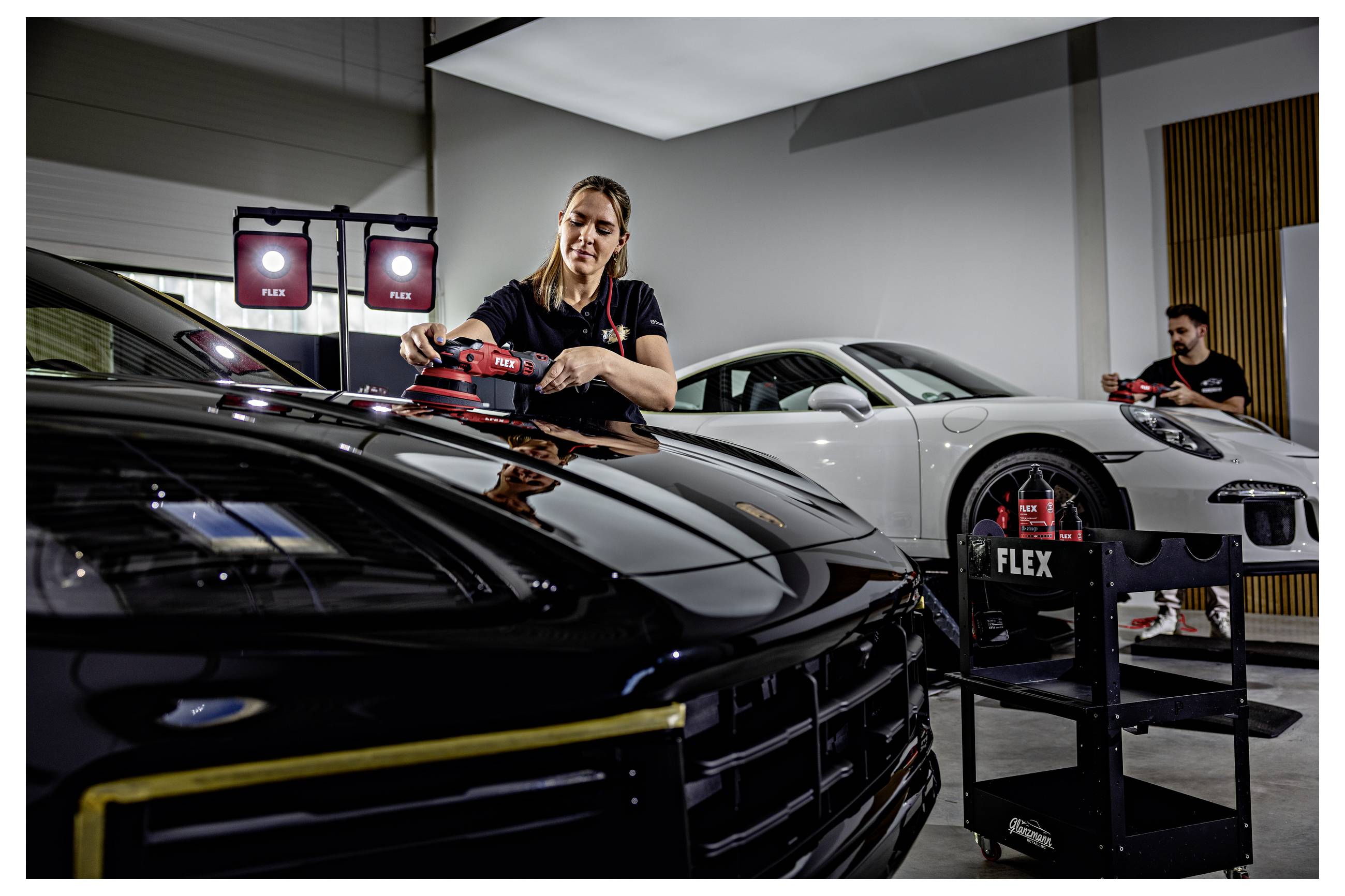 Detailer leští černý sportovní vůz v garáži, v pozadí bílý Porsche a přenosná světla.