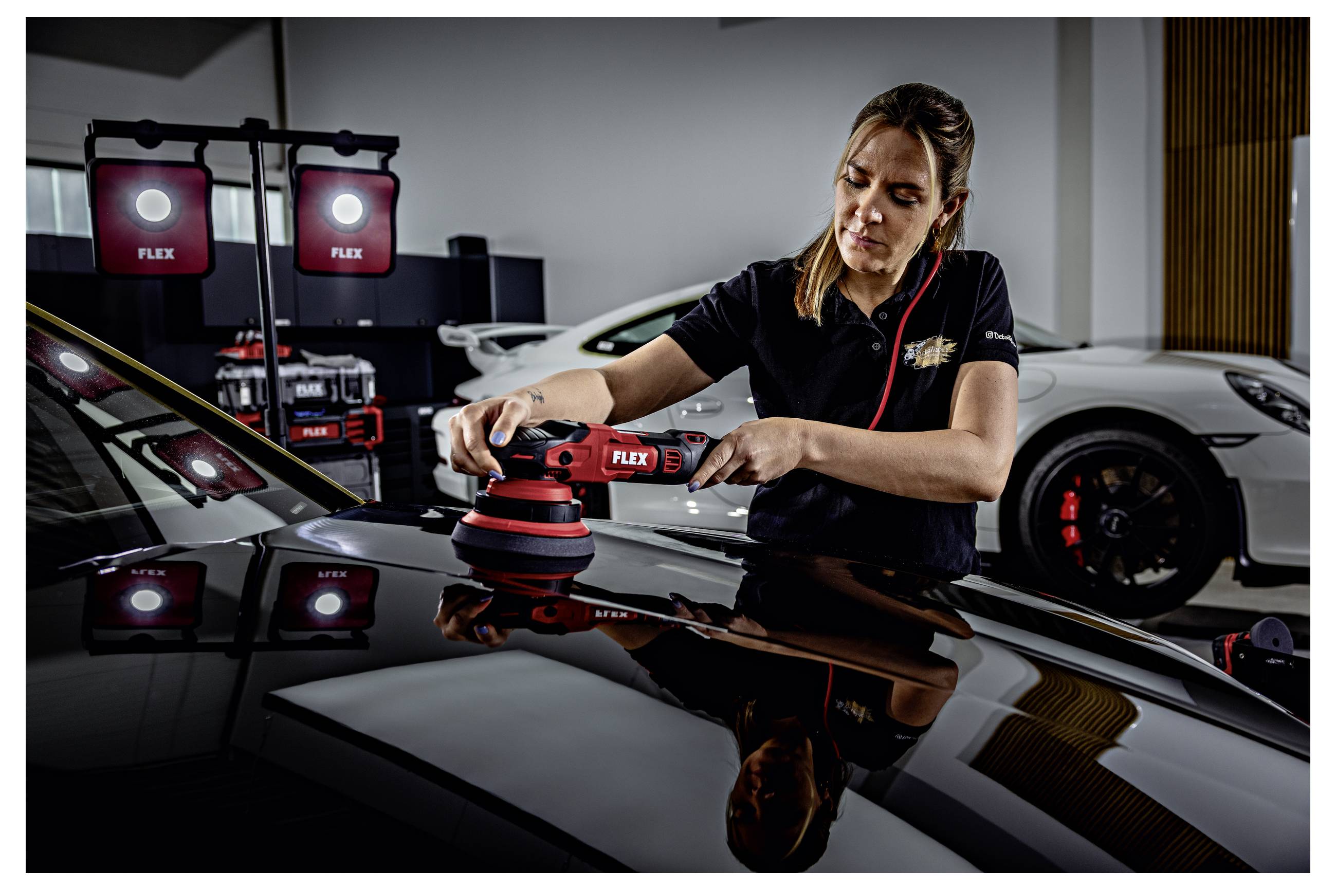 Technik leštící kapotu automobilu s FLEX leštičkou v detailingové dílně, se studiovými světly a sportovním autem v pozadí.