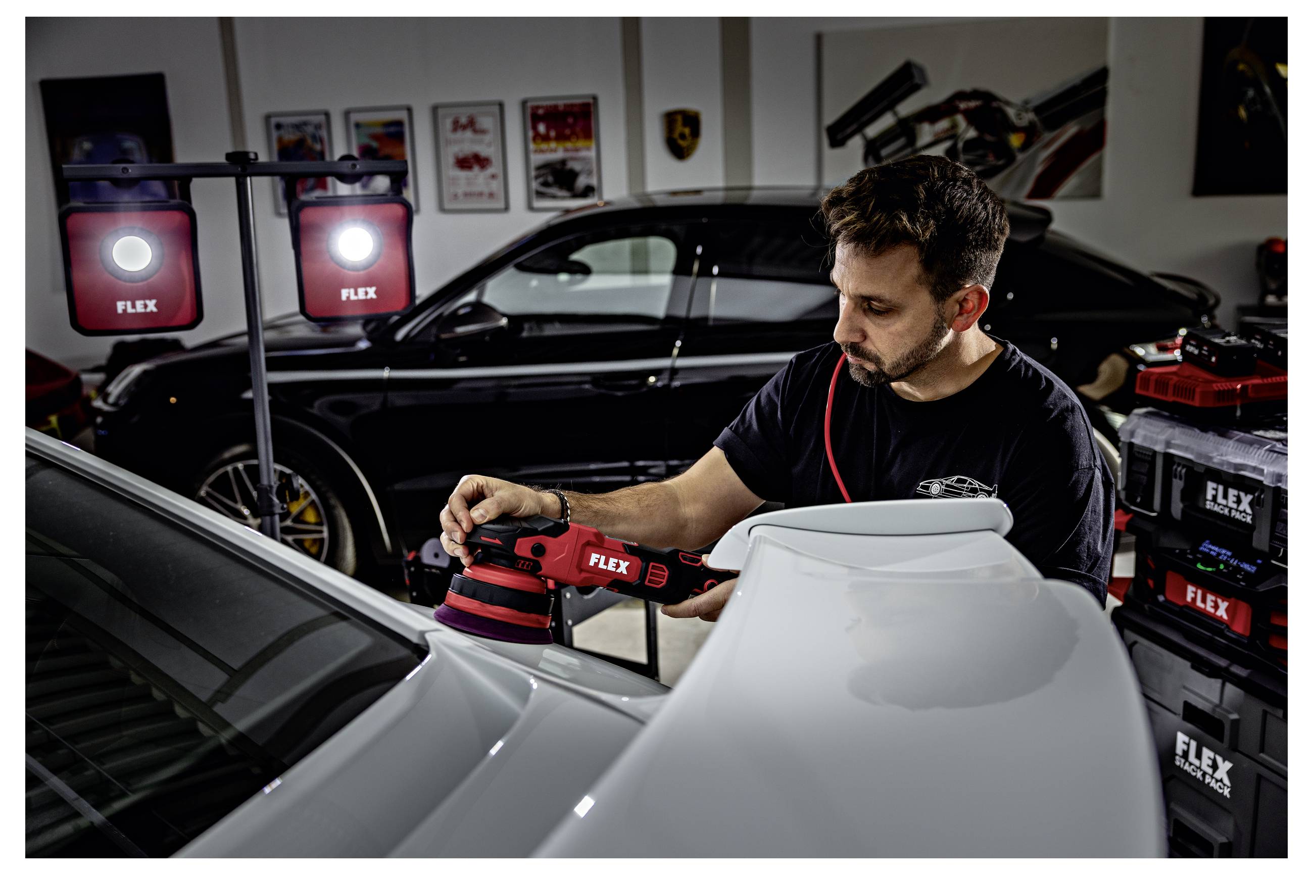 Muž leštící bílé auto v garáži pomocí FLEX leštičky, stojící vedle černého auta a značkových pracovních světel.